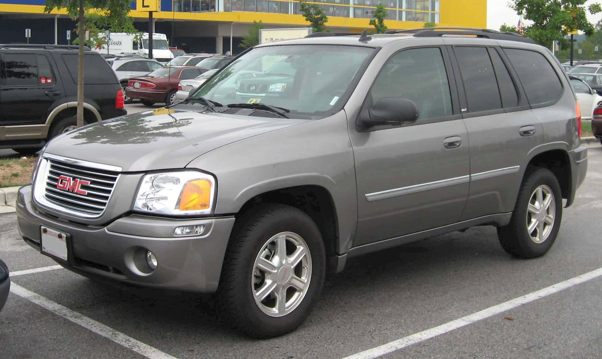 Gmc Envoy 桌布 壁紙 — 一輛灰色GMC Envoy SUV停放在陽光明媚的戶外停車場，背景有其他車輛和一棟黃色建築物。