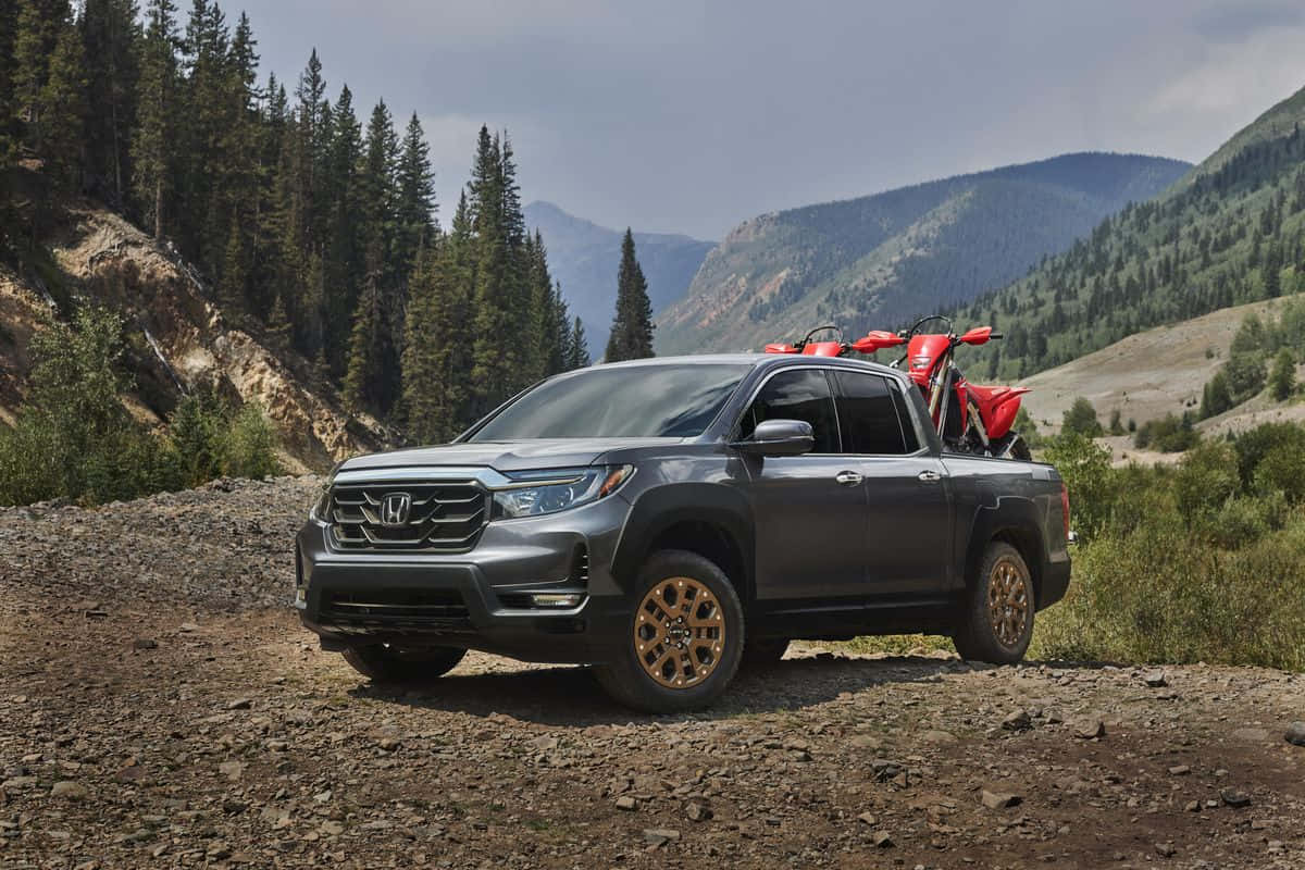 Honda Ridgeline 桌布 壁紙 — 一輛灰色本田Ridgeline皮卡車，帶有金色輪轂，車斗上載著一輛紅色電單車，停泊在岩石山徑上，周圍是常綠森林和遠處的山丘，天空多雲。