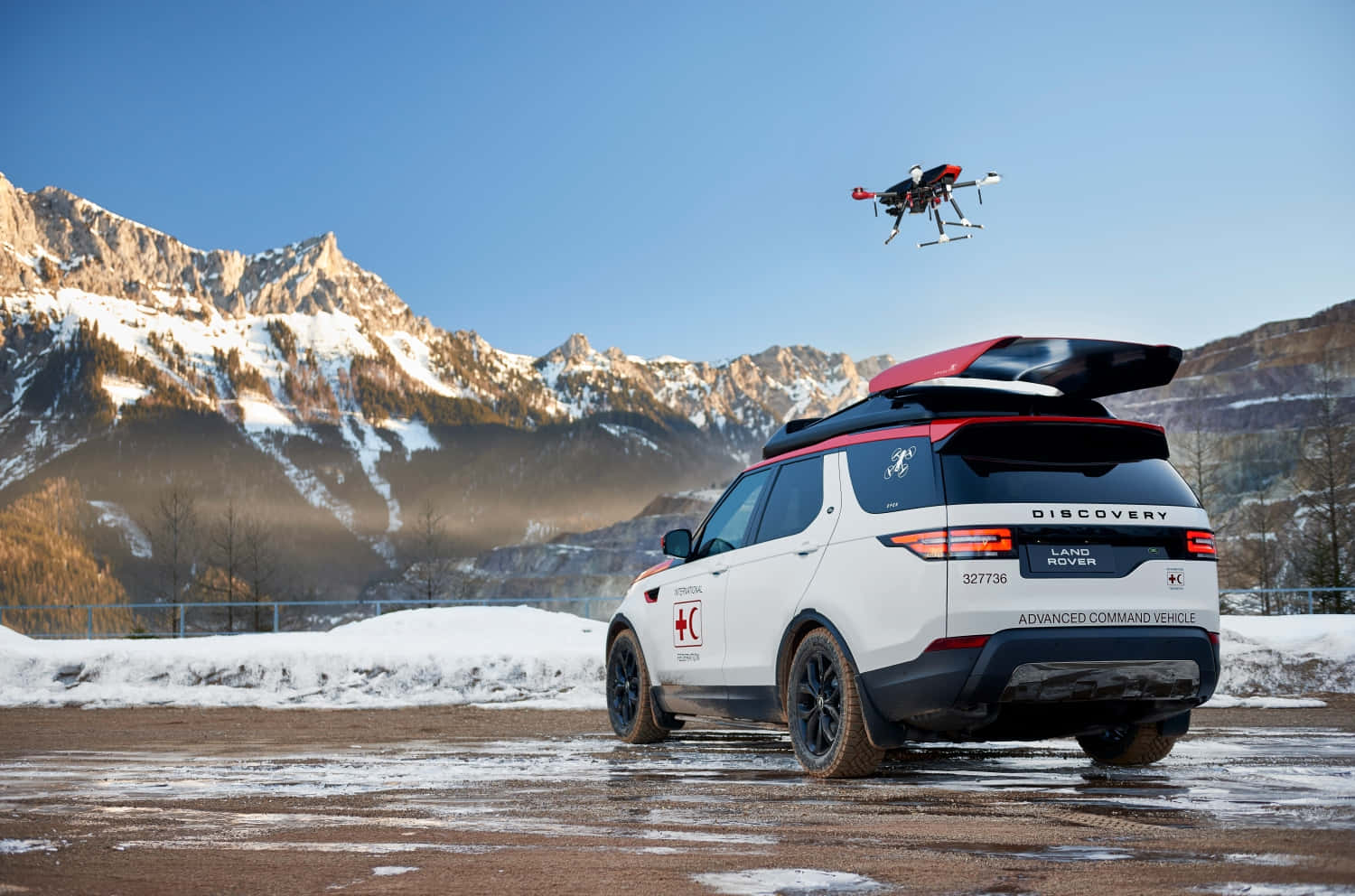 Land Rover Discovery 桌布 壁紙 — 一輛白色 Land Rover Discovery Advanced Command Vehicle，配有紅色車頂行李架和在其上方盤旋的無人機，停泊在積雪的山路上。