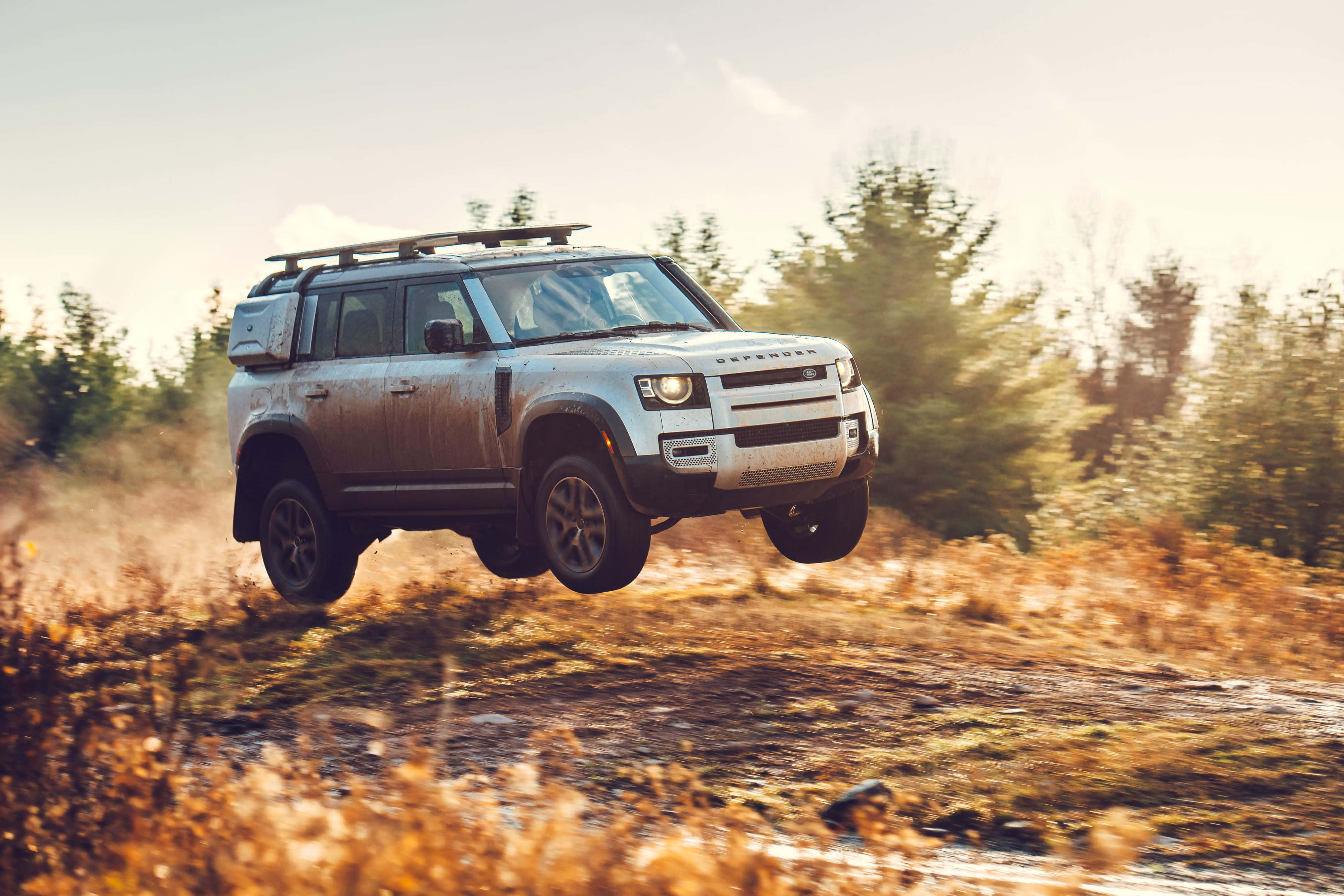 Land Rover Defender 桌布 壁紙 — 一輛白色 Land Rover Defender 在溫暖陽光下，背景有樹木，躍過金色草地。