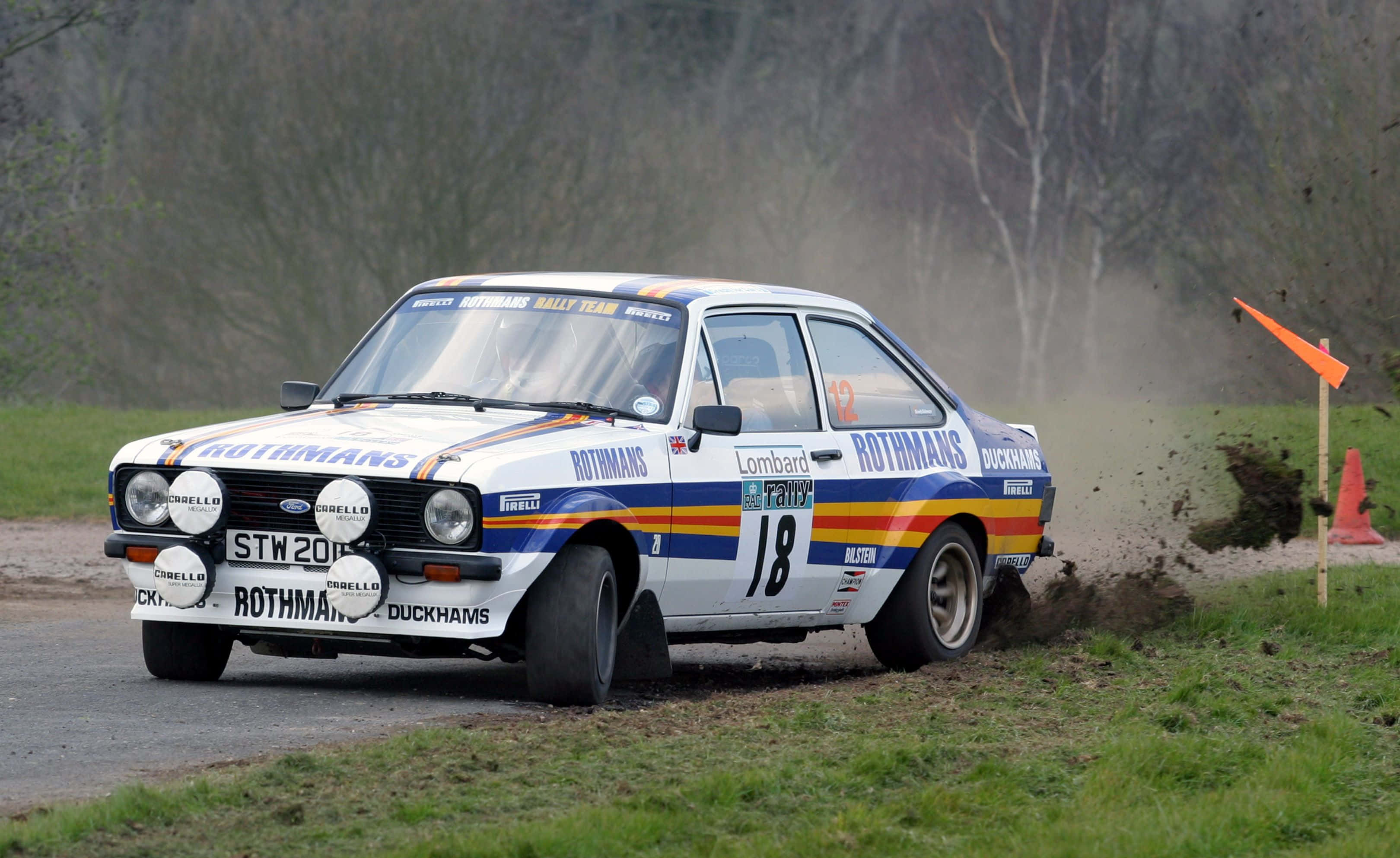 Ford Escort 桌布 壁紙 — 一輛白色 Rothmans 拉力賽車在標有橙色旗幟的草地森林賽道上疾馳，揚起塵土。