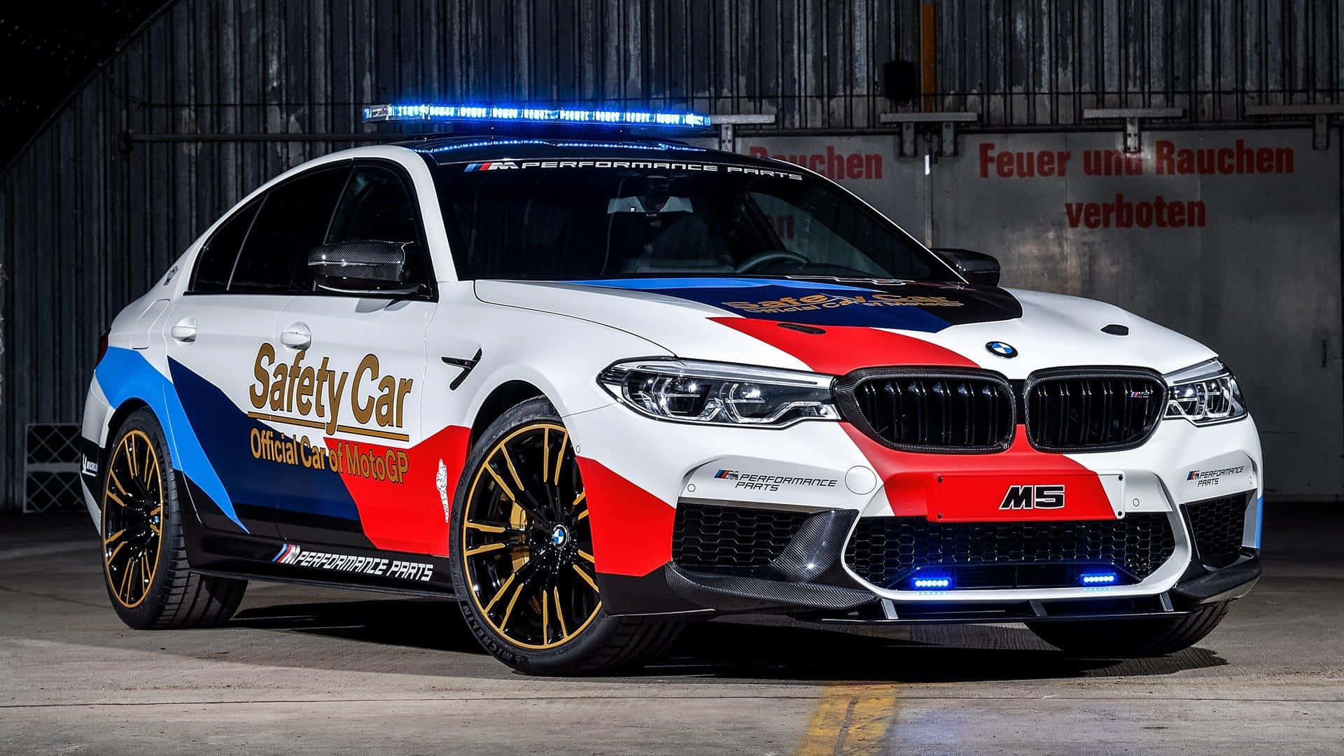 Safety Car 桌布 壁紙 — 一輛白色、紅色和藍色的BMW M5安全車，閃爍著燈光，停在昏暗的工業車庫中。
