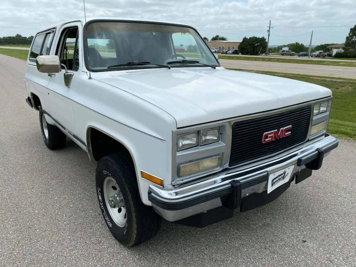 Gmc Jimmy 桌布 壁紙 — 一輛白色1980年代GMC Jimmy SUV停泊在碎石地上，天空多雲，背景是郊區建築物。