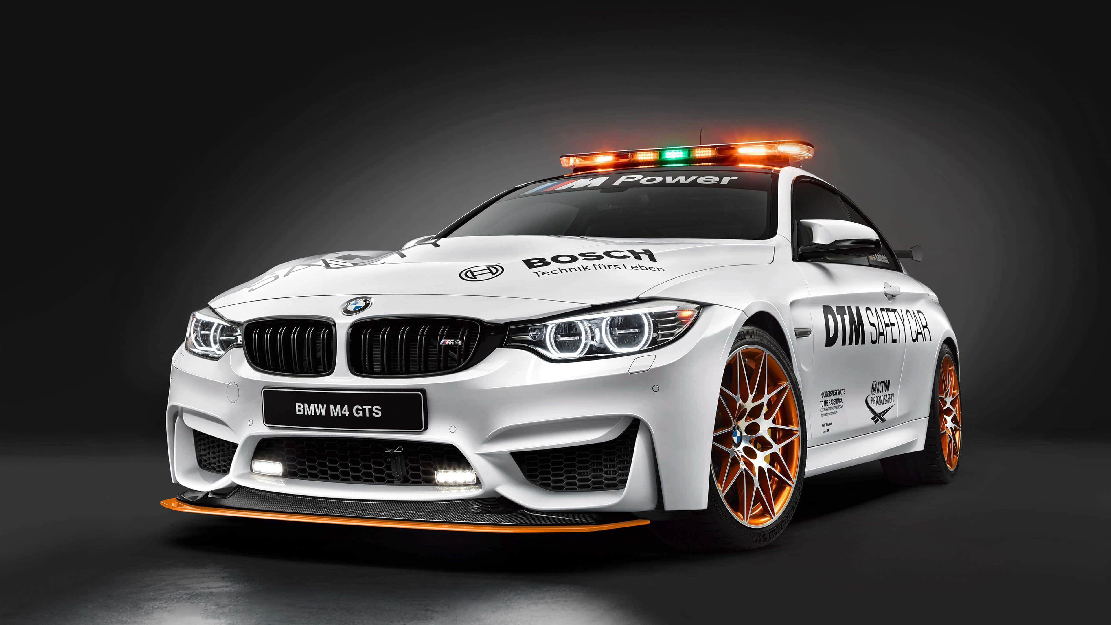 Safety Car 桌布 壁紙 — 一輛白色BMW M4 GTS安全車，帶有橙色飾邊和閃爍燈光，置於深色工作室背景前。