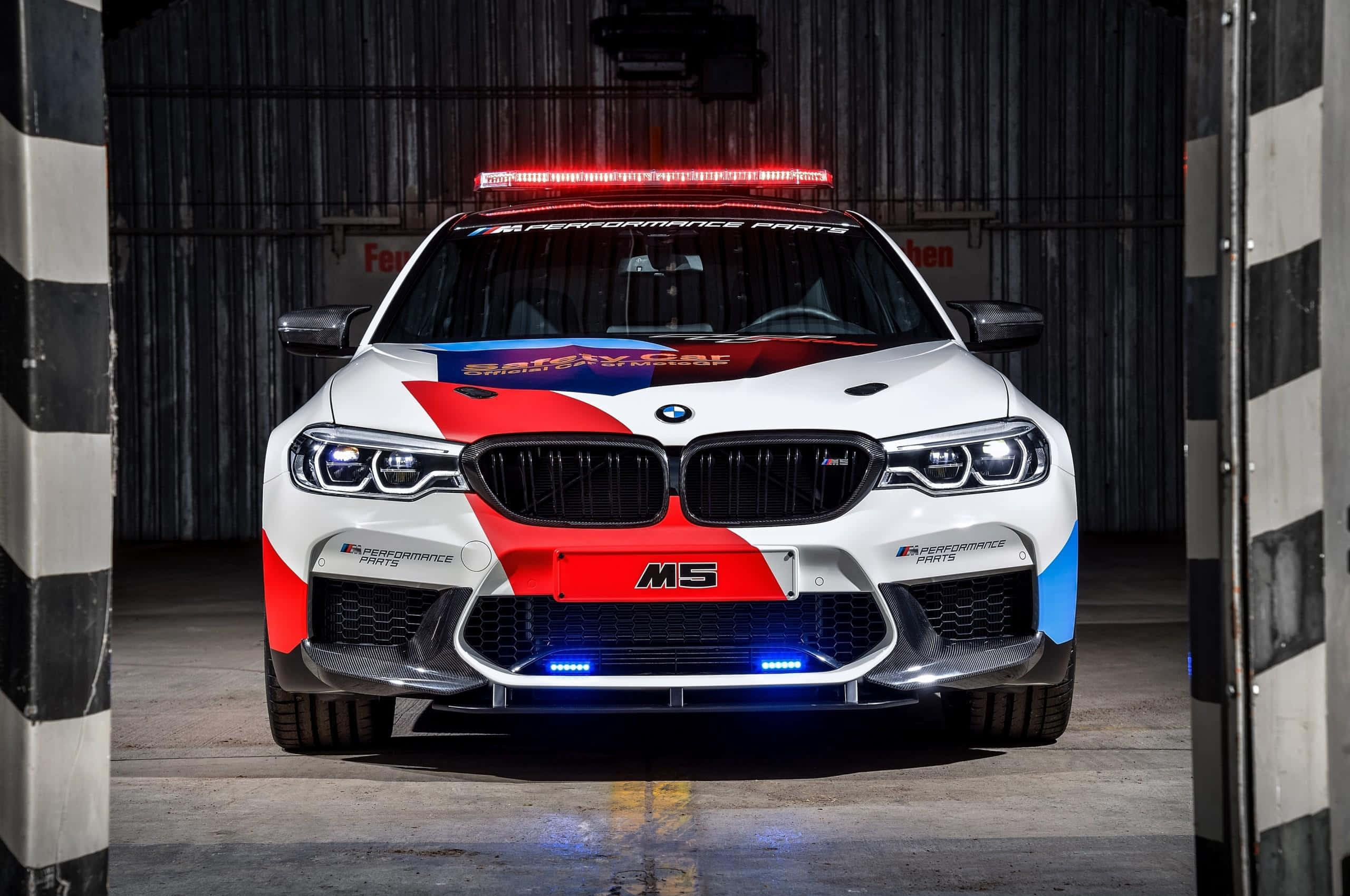 高性能安全車在賽道上奔馳 壁紙 — 一輛白色BMW M5安全車，帶有紅藍色塗裝和閃爍燈光，停放在由黑白條紋柱子框起的車庫中。