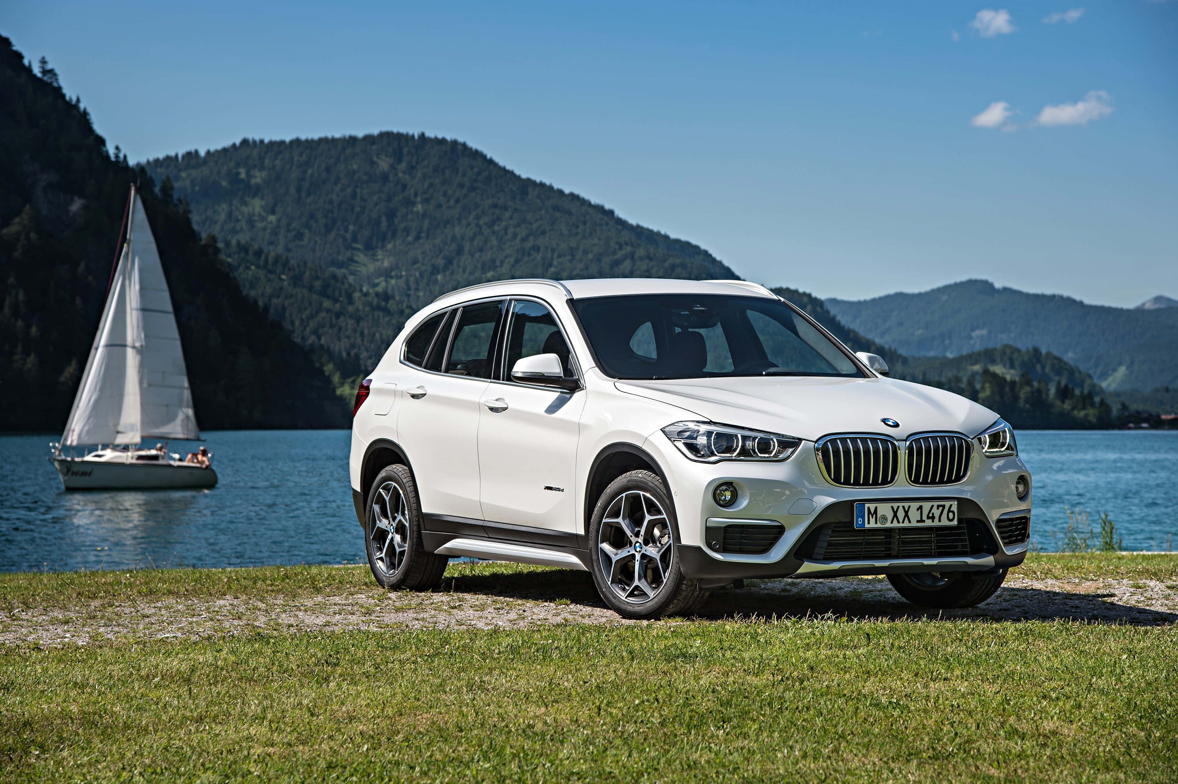 Bmw X1 桌布 壁紙 — 一輛白色BMW SUV停泊在湖邊草地上，湖上有帆船和樹木繁茂的山丘，天空湛藍清澈。