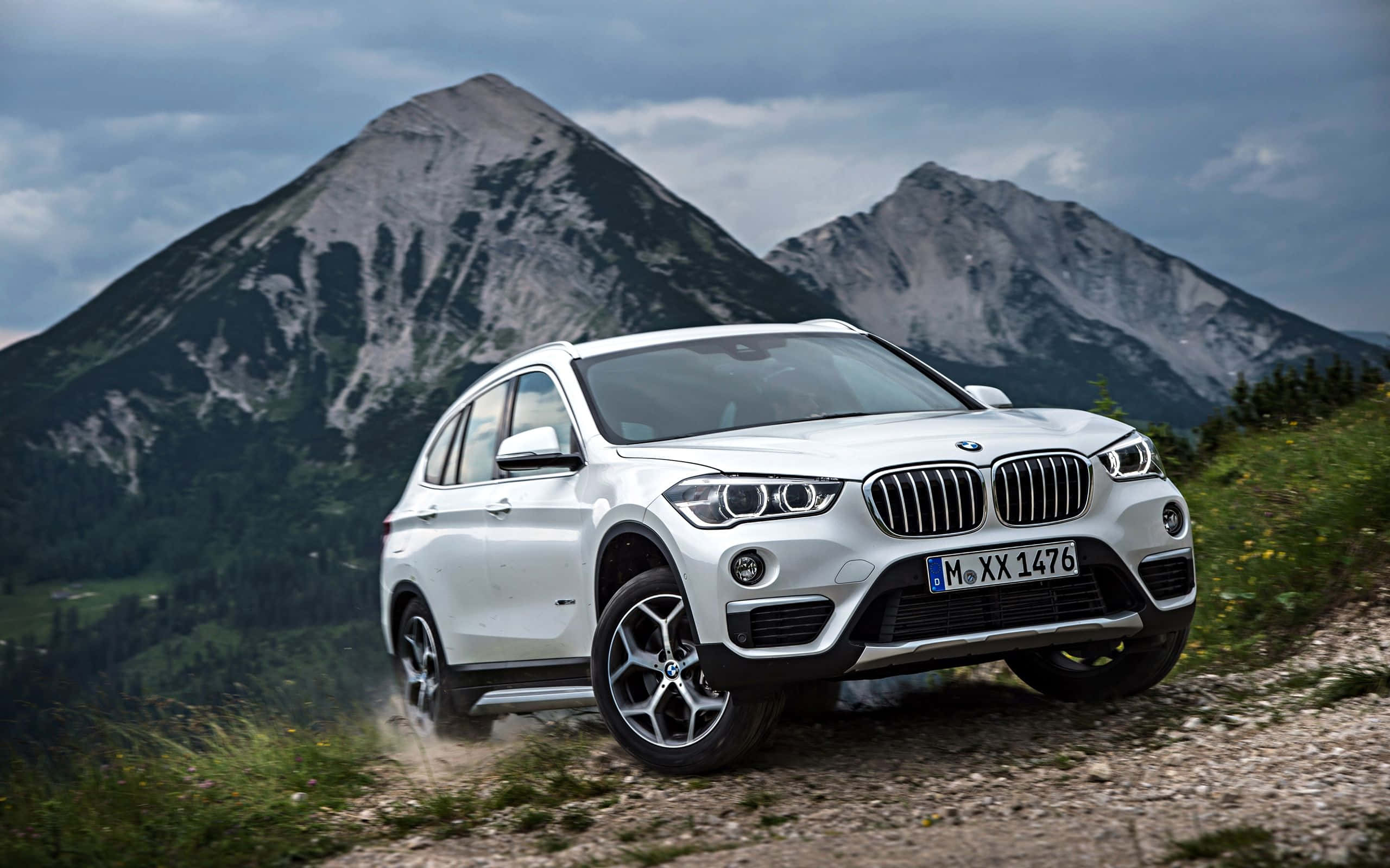 Bmw X1 桌布 壁紙 — 一輛白色BMW SUV在碎石路上行駛，背後有崎嶇的山脈在陰天下聳立。