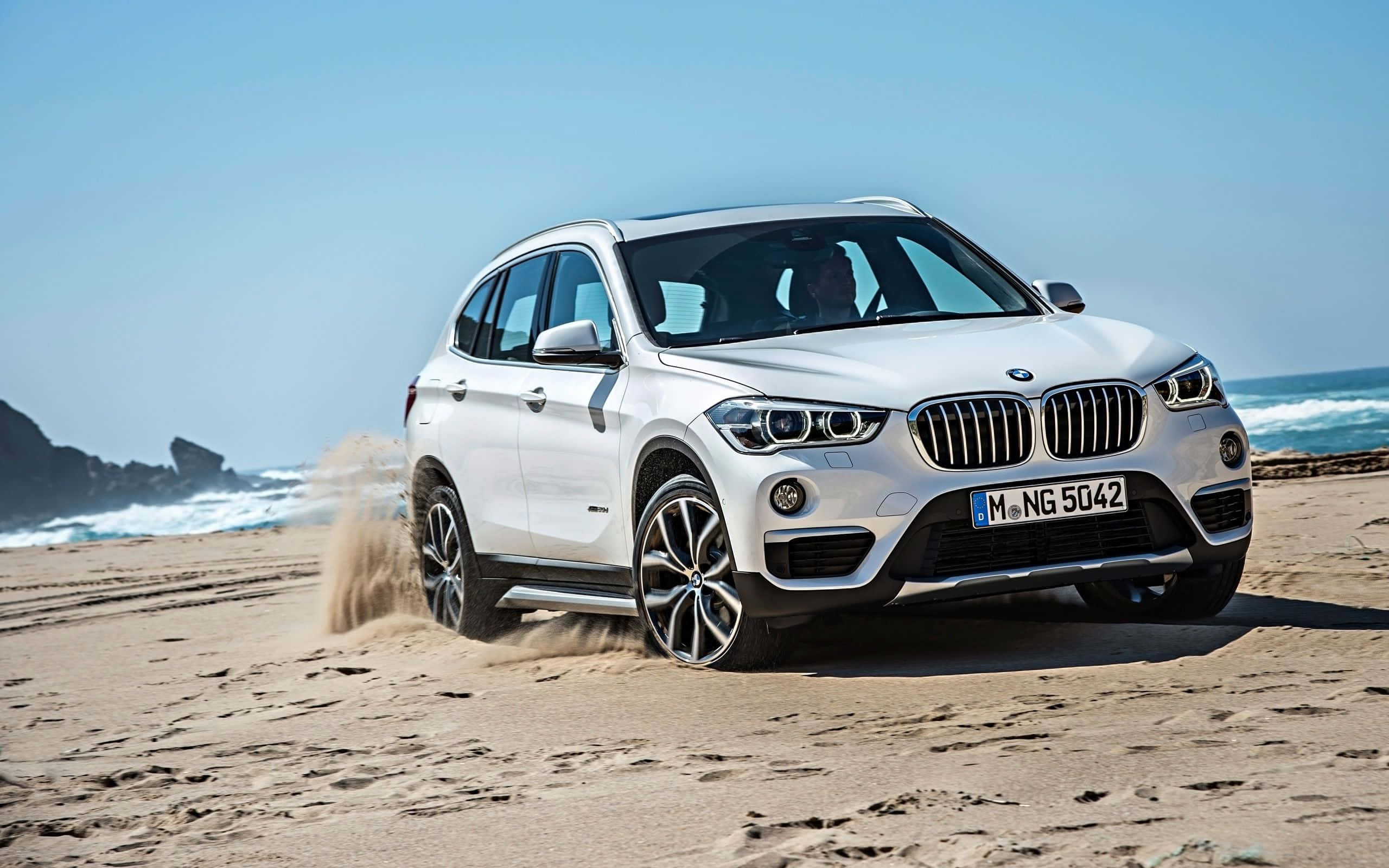 Bmw X1 桌布 壁紙 — 一輛白色BMW SUV在陽光明媚的海灘上行駛，背景是海浪和岩石峭壁。