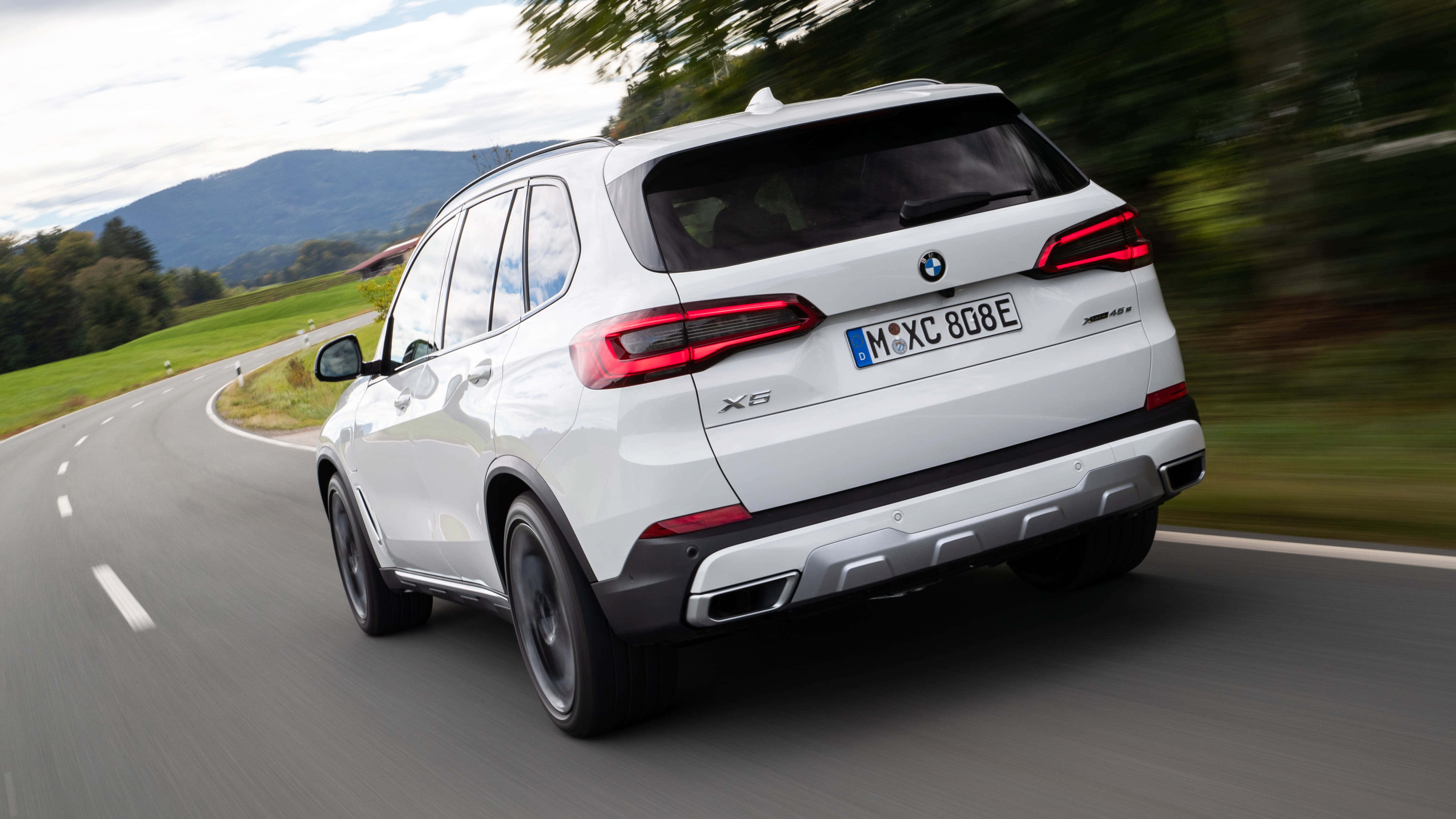 Bmw X5 桌布 壁紙 — 一輛白色BMW X5 SUV在蜿蜒的山路上疾馳，背景是模糊的綠意和遠處的山丘，天空部分多雲。