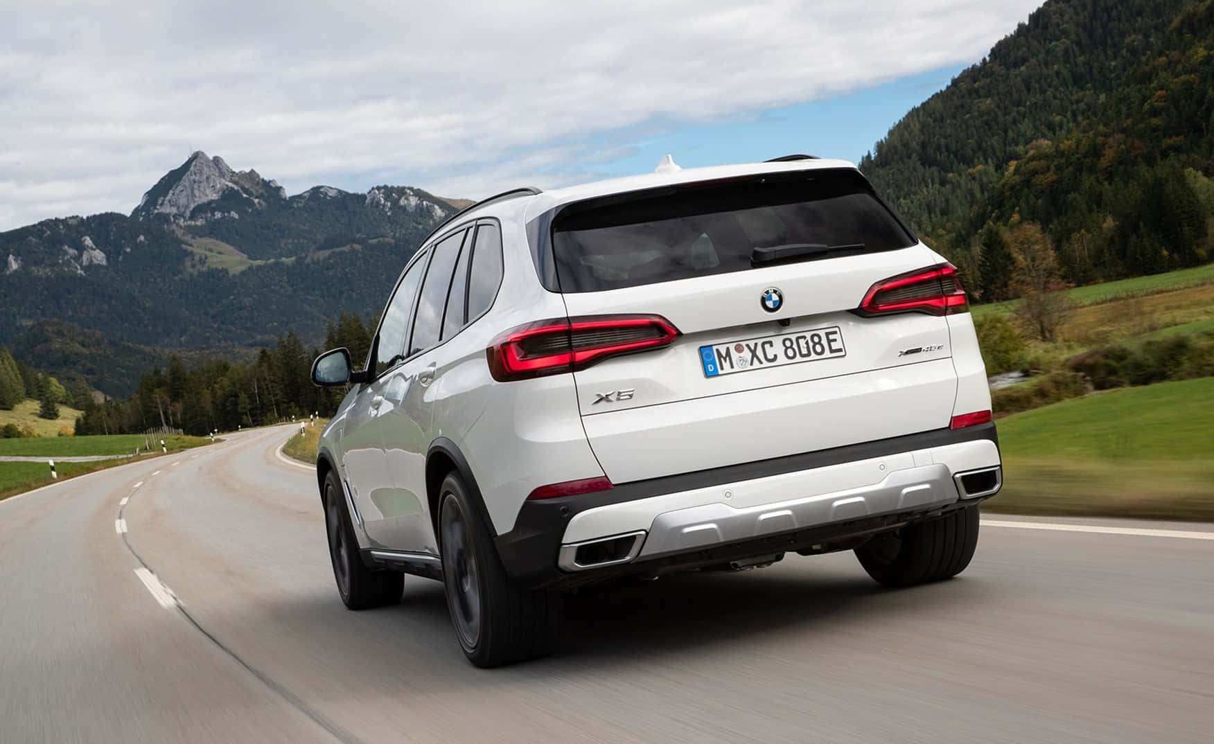 Bmw X5 桌布 壁紙 — 一輛白色BMW X5 SUV沿著蜿蜒的山路行駛，穿過森林覆蓋的山丘和岩石山峰，天空多雲。