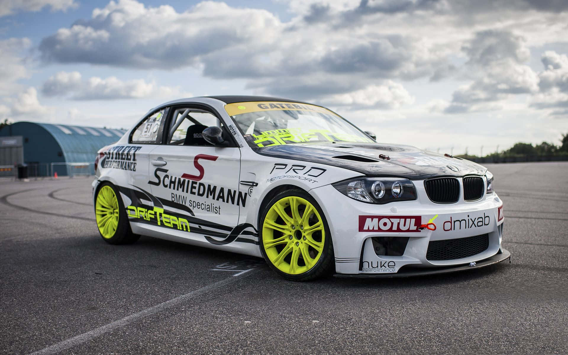 Bmw 1 Series 桌布 壁紙 — 一輛白色BMW飄移車配備螢光黃色輪胎，在多雲的天空下於瀝青賽道上奔馳。