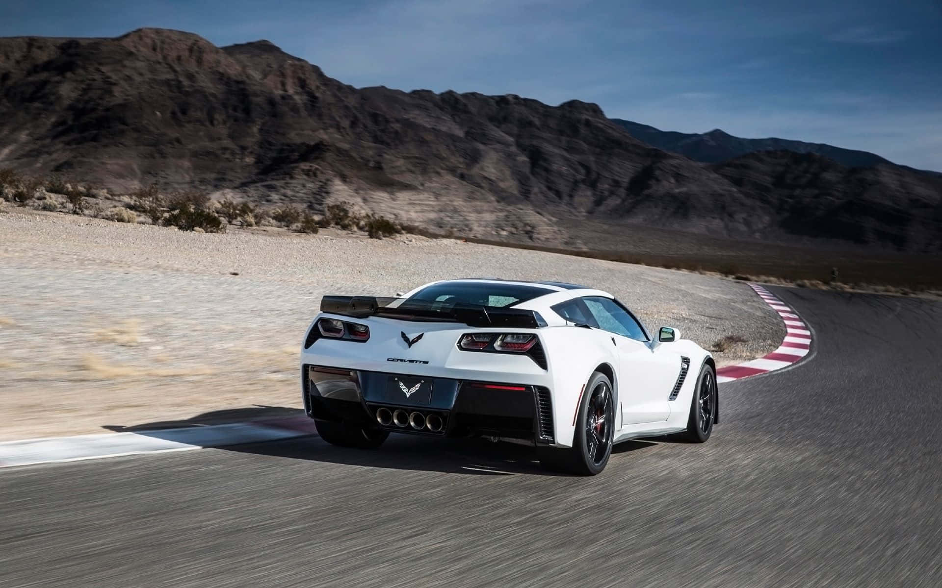 Chevrolet Corvette Z06 桌布 壁紙 — 一輛白色Corvette C7 Stingray在沙漠賽道上疾馳，賽道有紅色和白色的路緣石，背景是晴朗藍天下巍峨的山脈。