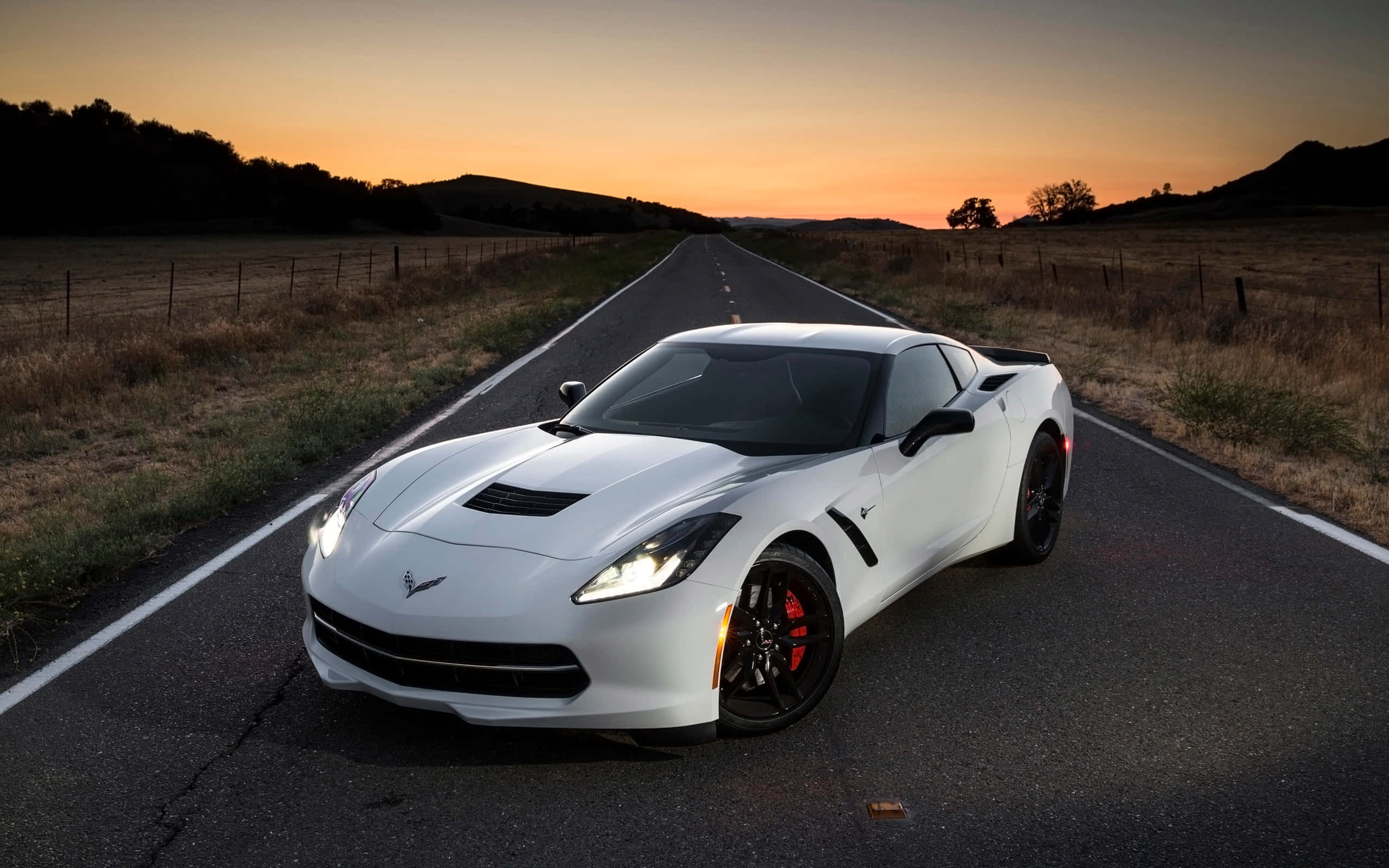 Chevrolet Corvette Stingray 桌布 壁紙 — 一輛白色Corvette跑車在日落時分的長長鄉村道路上閃耀，伴隨著溫暖的橙色天空和剪影般的山丘。