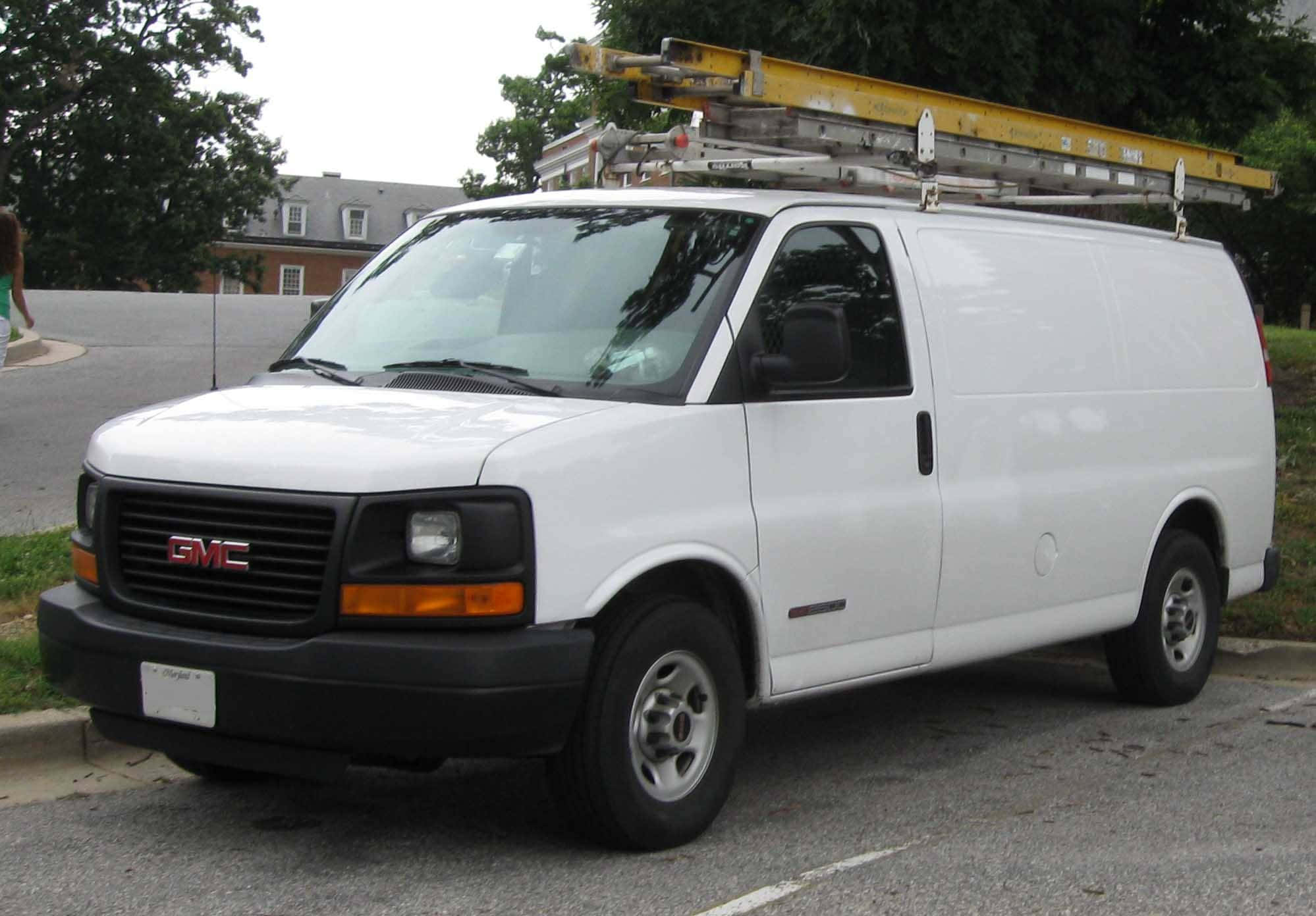 Gmc Savana 桌布 壁紙 — 一輛白色GMC客貨車，車頂裝有黃色梯子，停在路邊，旁邊有綠樹。