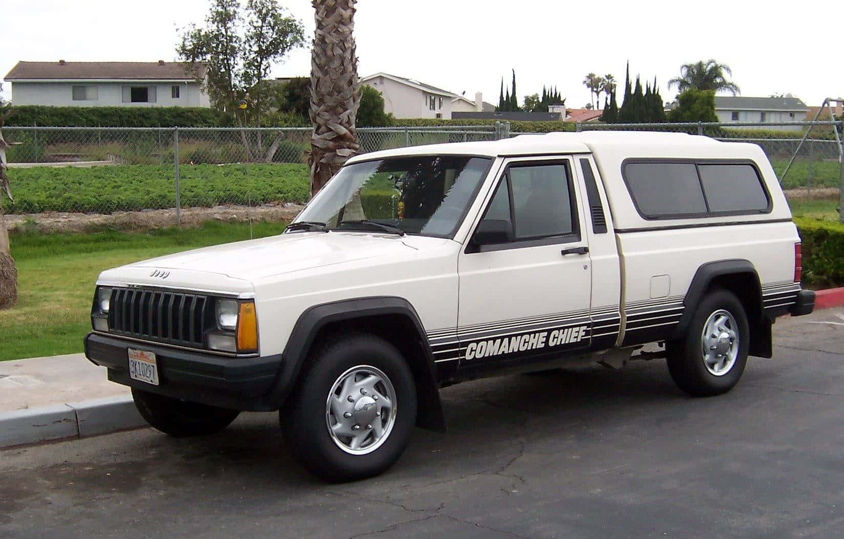 Jeep Comanche 桌布 壁紙 — 一輛白色Jeep Cherokee Comanche Chief旅行車停泊在一個裂開的瀝青停車場，背景有棕櫚樹和郊區房屋。