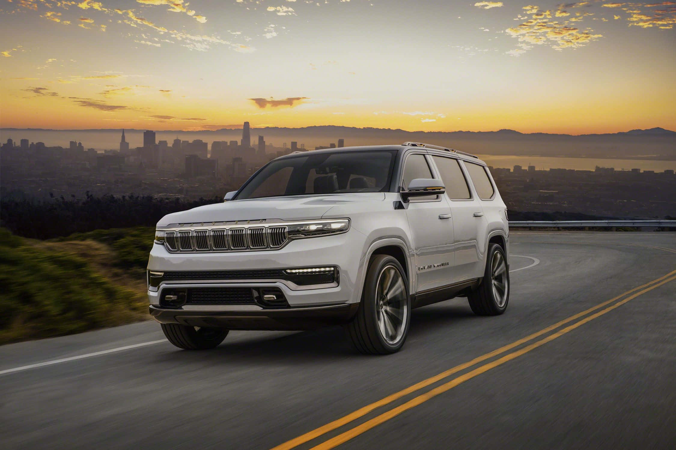 Jeep Wagoneer 桌布 壁紙 — 一輛白色Jeep Grand Wagoneer SUV在日落時分行駛在蜿蜒的道路上，背景是城市天際線和山脈。