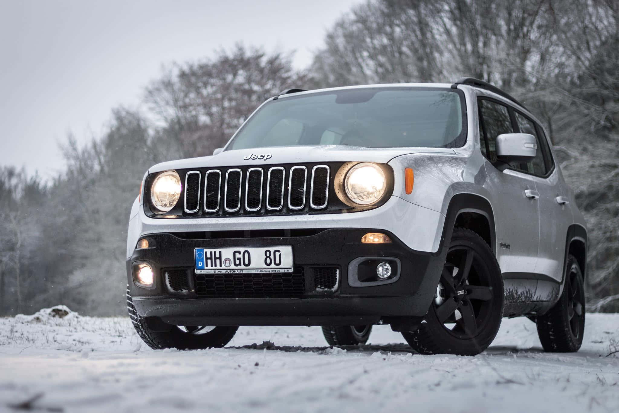 Jeep Renegade 桌布 壁紙 — 一輛白色Jeep Renegade SUV亮著車頭燈，停泊在雪地上，四周是光禿禿的冬季樹木，天空多雲。