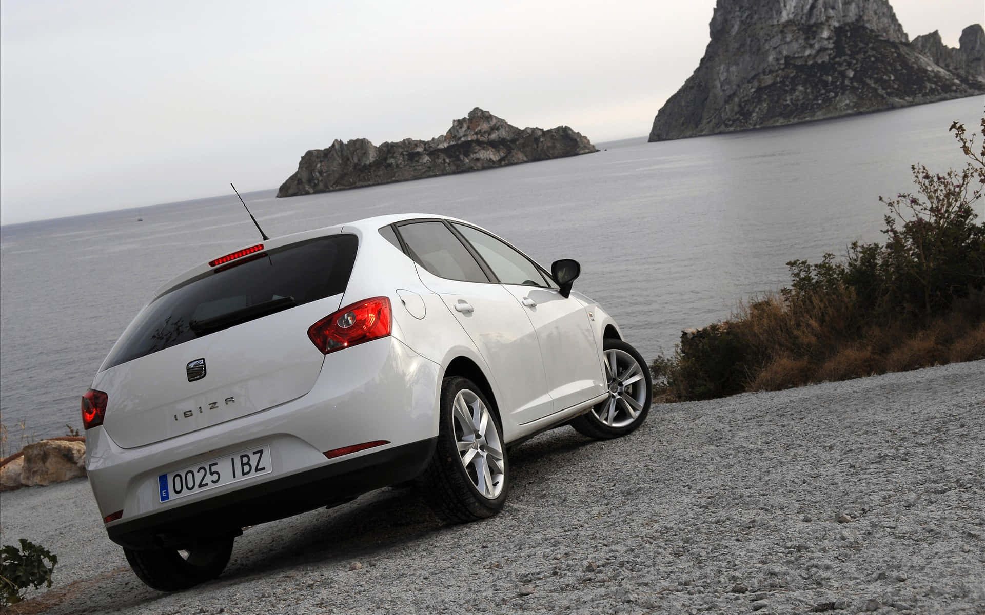 seat 桌布 壁紙 — 一輛白色Seat Ibiza停泊在碎石懸崖上，俯瞰著平靜的海面，遠處有岩石島嶼，天空多雲。