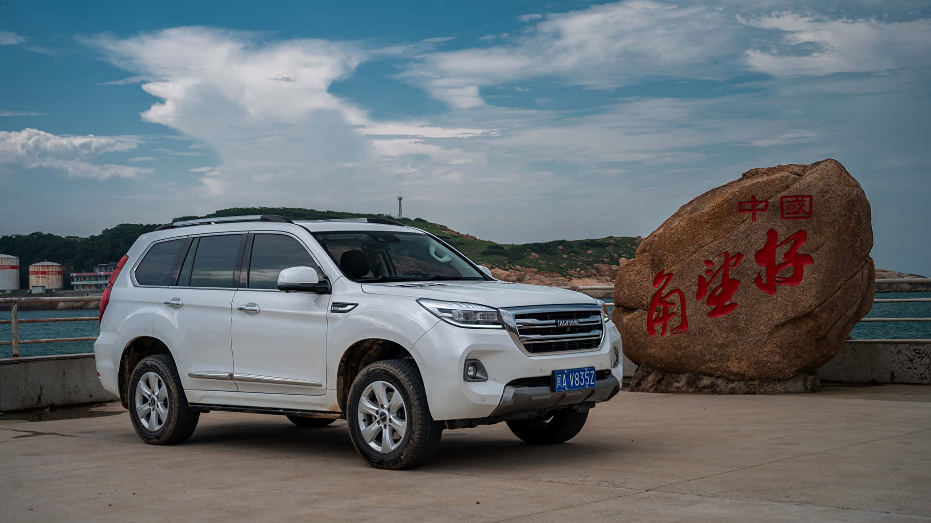 Haval 桌布 壁紙 — 一輛白色SUV停泊在海邊一塊刻有文字的大石旁，天空部分多雲。