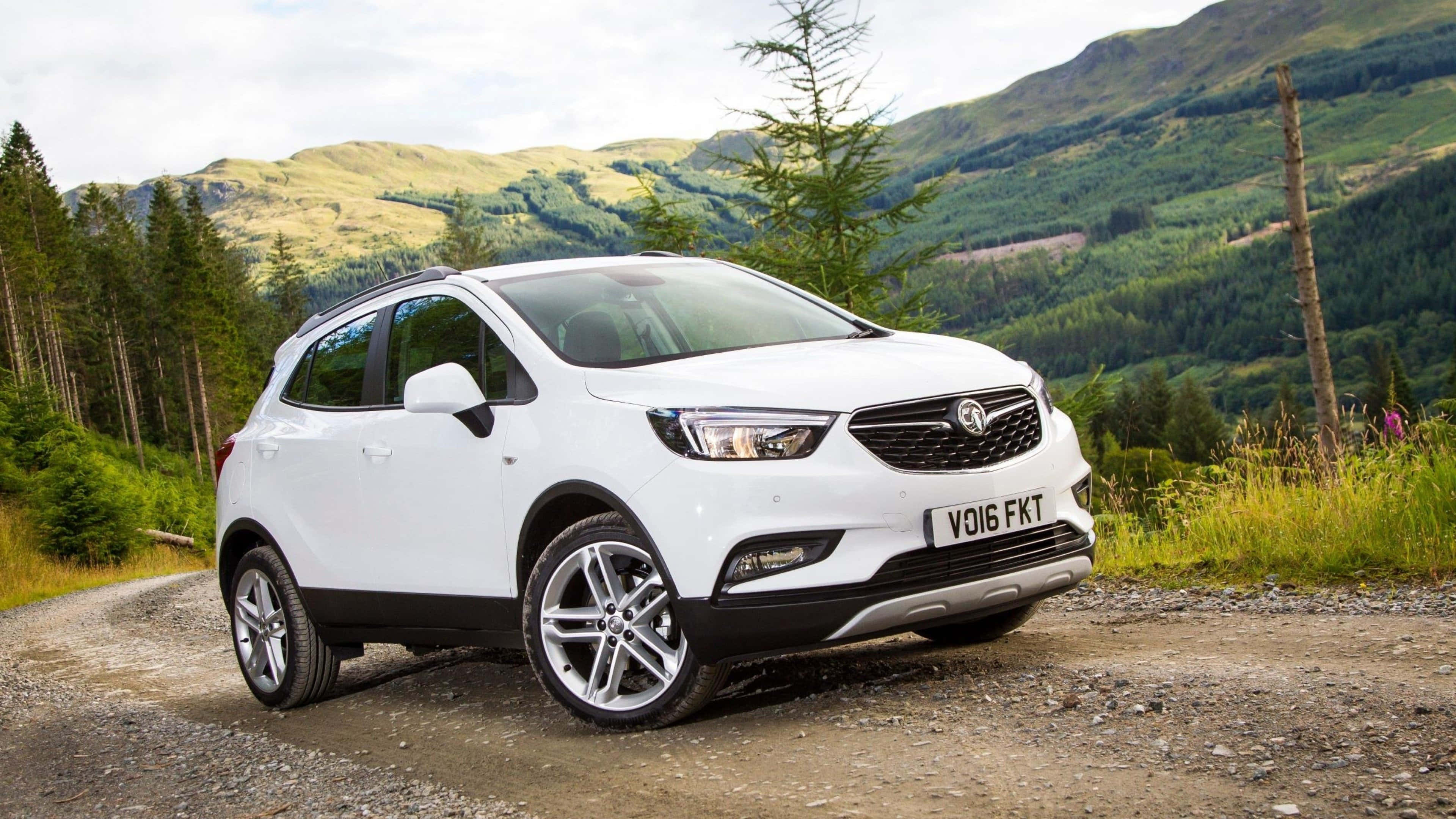Opel Mokka 桌布 壁紙 — 一輛白色SUV停泊在碎石路上，背景是起伏的綠色山丘和松樹。