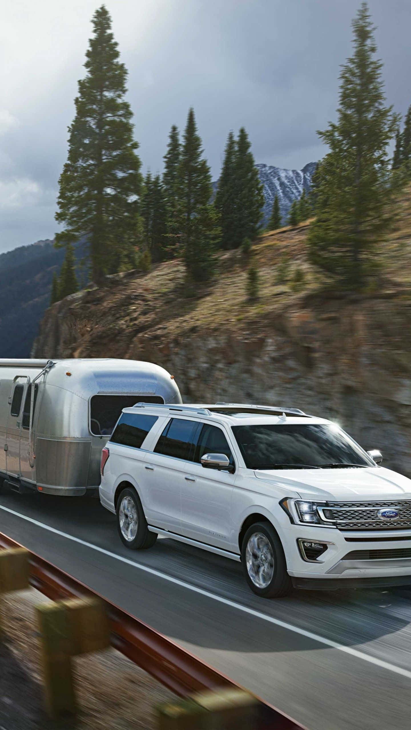 Ford Expedition 桌布 壁紙 — 一輛白色SUV拖著銀色Airstream拖車，在兩旁高聳松樹和積雪山峰環繞的山路上疾馳。