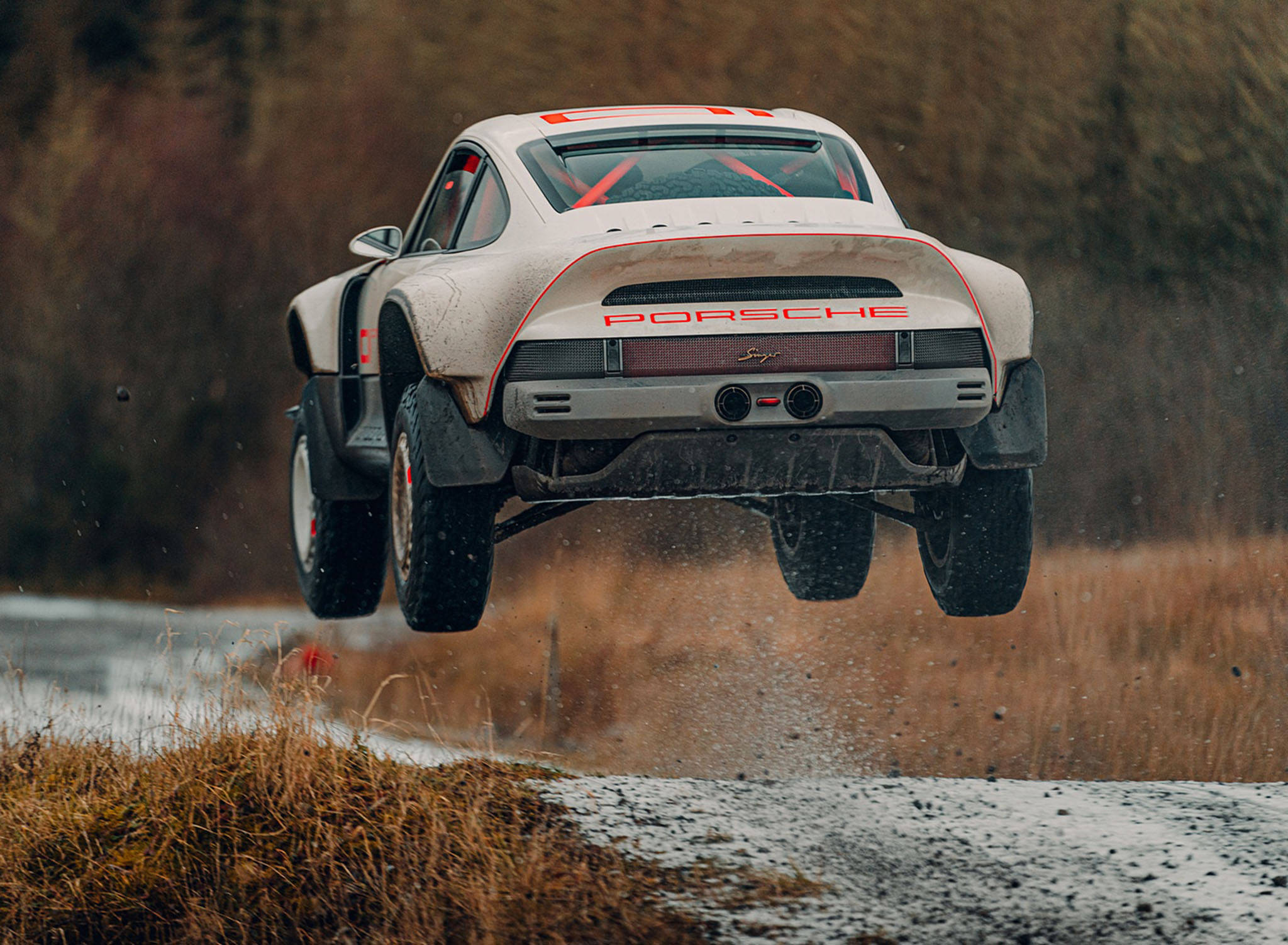 Singer Porsche Safari 911 壁紙 — 一輛白色保時捷拉力賽車在泥濘賽道上空飛馳，背景是秋日樹木。