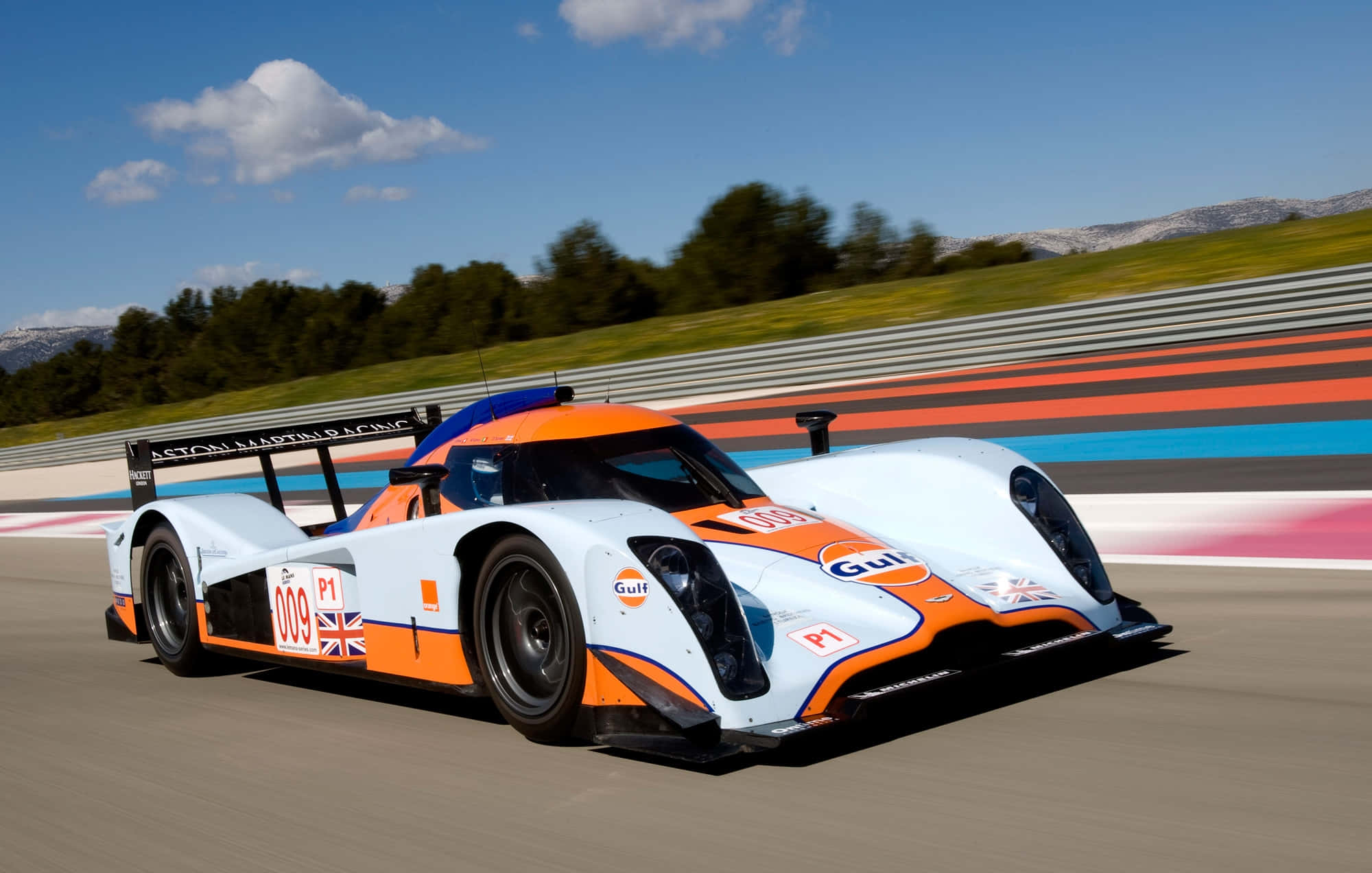 Car Racing 桌布 壁紙 — 一輛白色和橙色的Aston Martin賽車在明亮的藍天下，以模糊的動態沿著賽道飛馳。