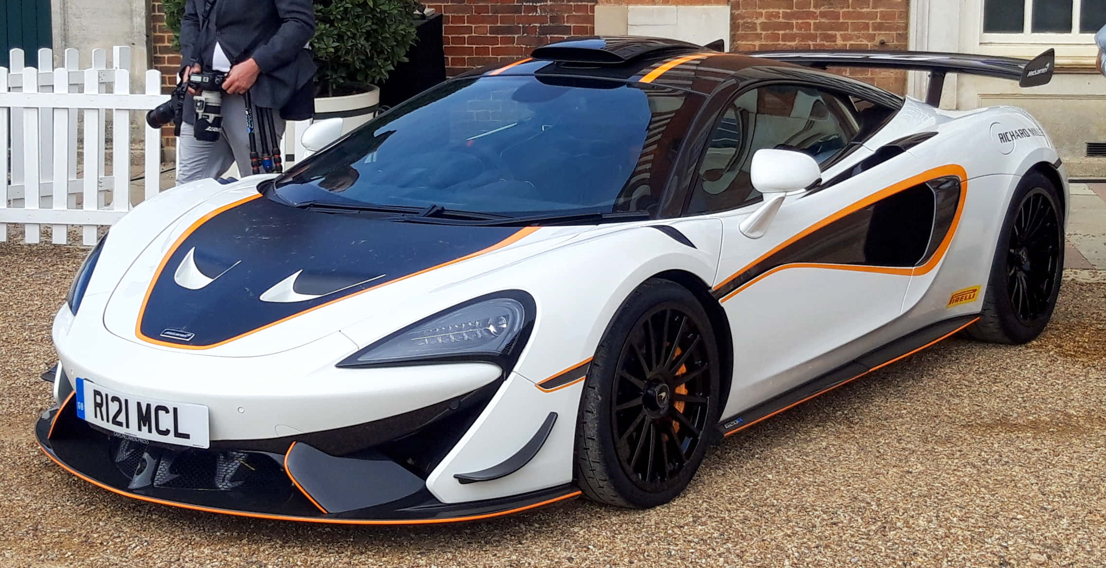 Mclaren 620r 桌布 壁紙 — 一輛白色和橙色的McLaren Senna超級跑車，帶有黑色點綴，停在磚砌建築前的礫石上。