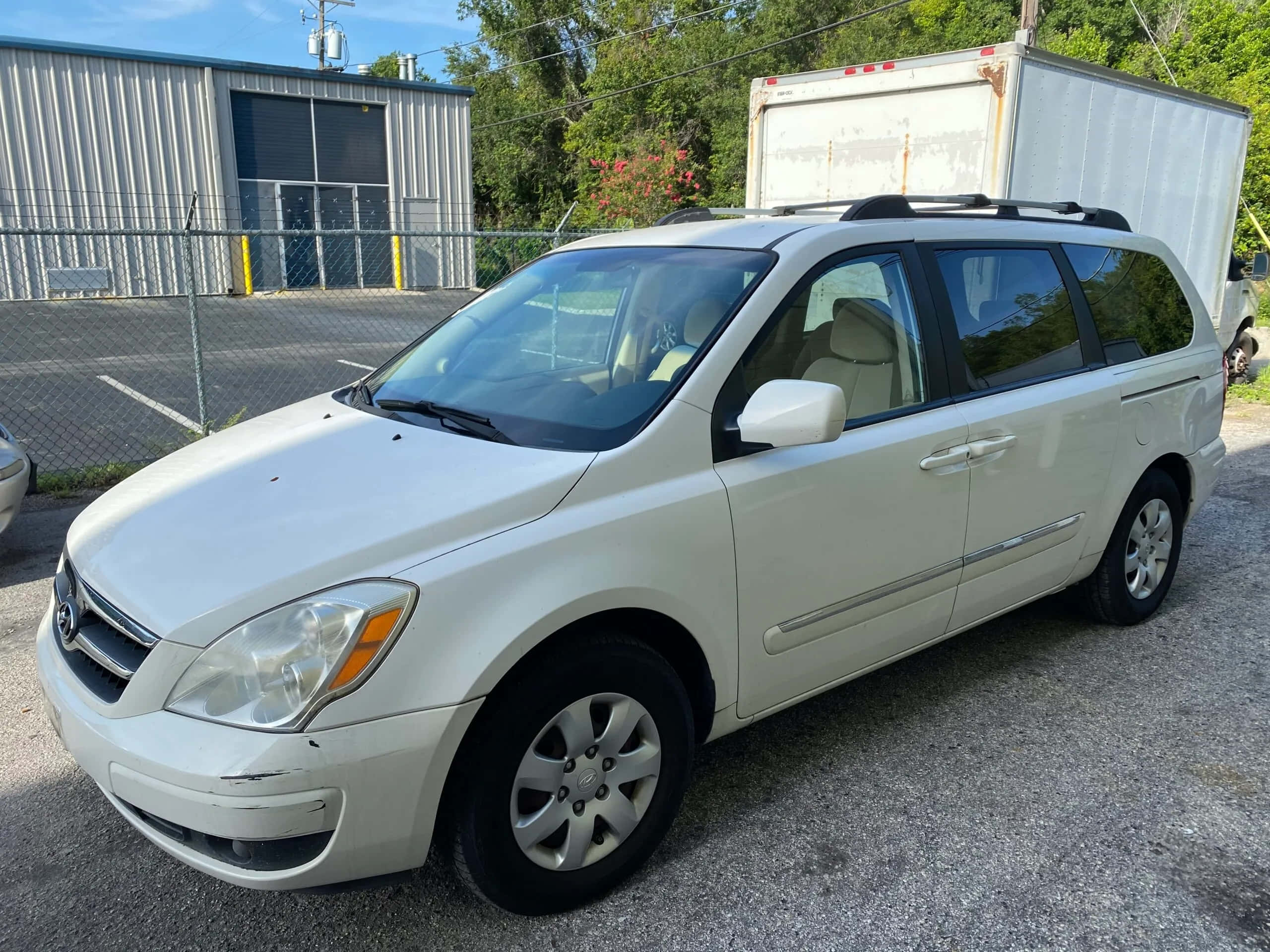 Hyundai Entourage 桌布 壁紙 — 一輛白色小型貨車停泊在碎石地上，背景有鐵絲網圍欄和倉庫，在明亮的日光下。