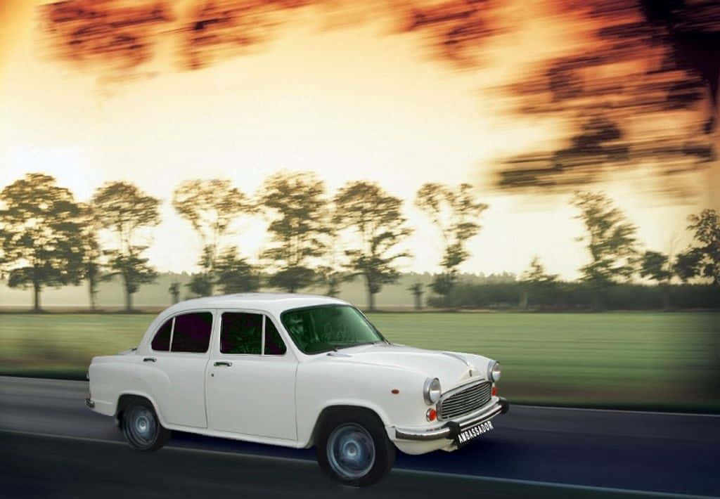 Hindustan Motors 桌布 壁紙 — 一輛白色復古汽車沿著道路疾馳，背景是模糊的樹木和移動中的金色日落天空。