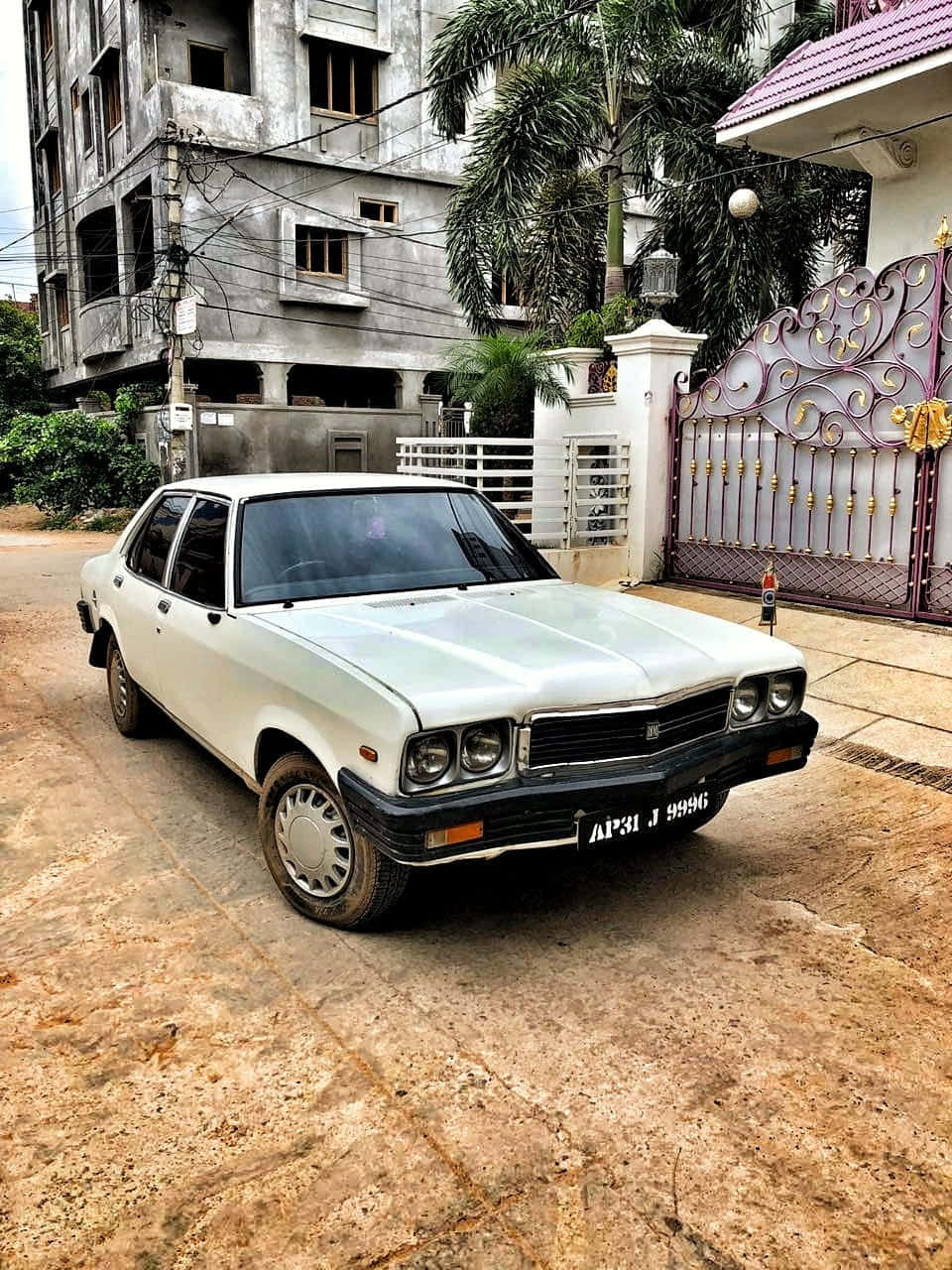 Hindustan Motors 桌布 壁紙 — 一輛白色復古轎車停泊在塵土飛揚的車道上，旁邊是華麗的大閘和未完成的混凝土建築。