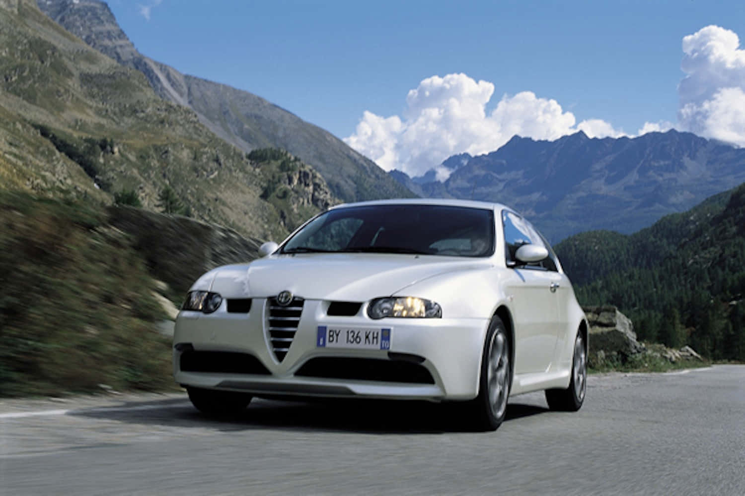 Alfa Romeo 147 桌布 壁紙 — 一輛白色愛快羅密歐跑車在晴朗藍天白雲下，沿著蜿蜒的山路飛馳。