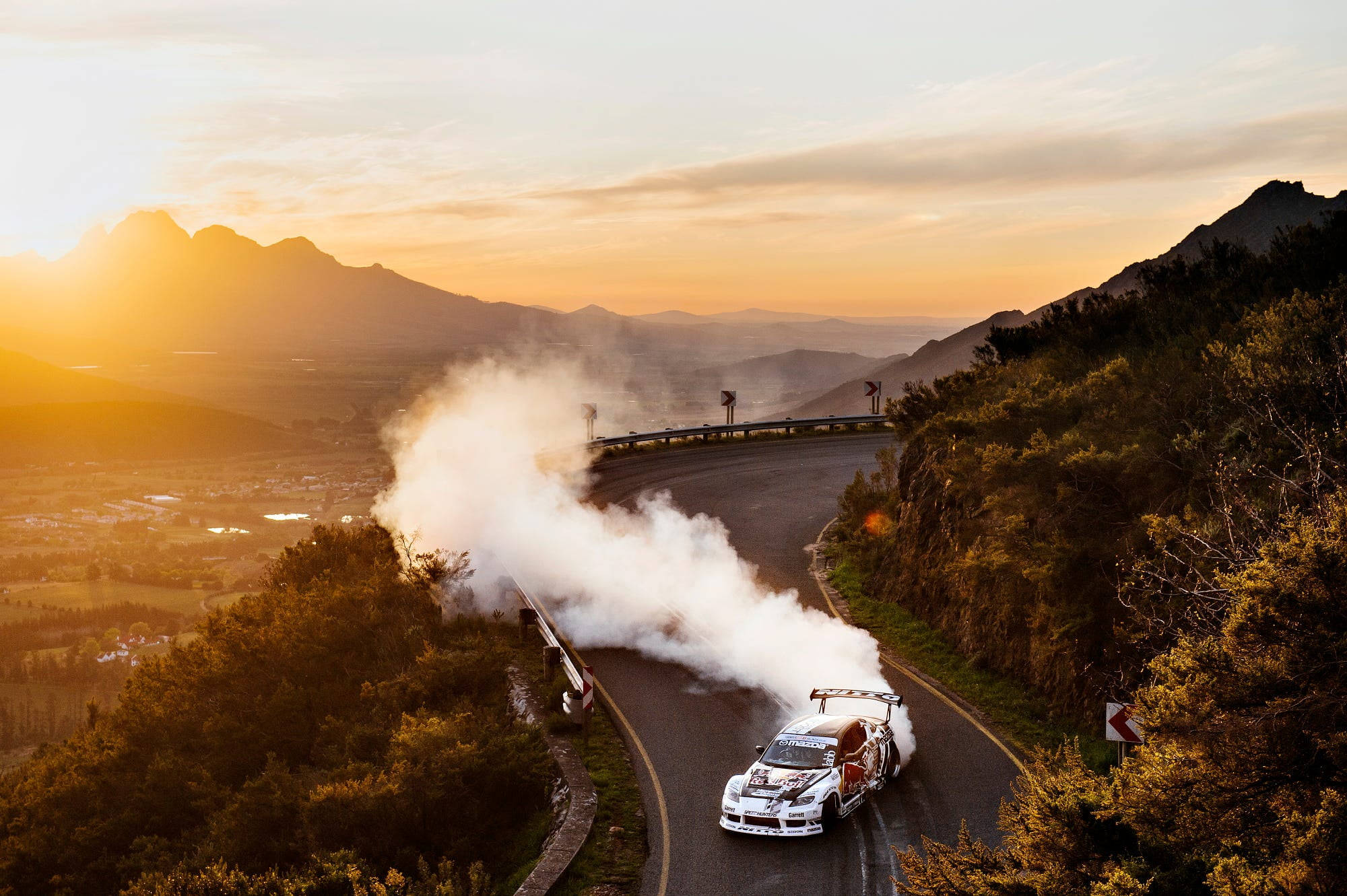 Mountain 桌布 壁紙 — 一輛白色拉力賽車在日落時分於山彎飄移，揚起濃煙，映襯著金色山丘。