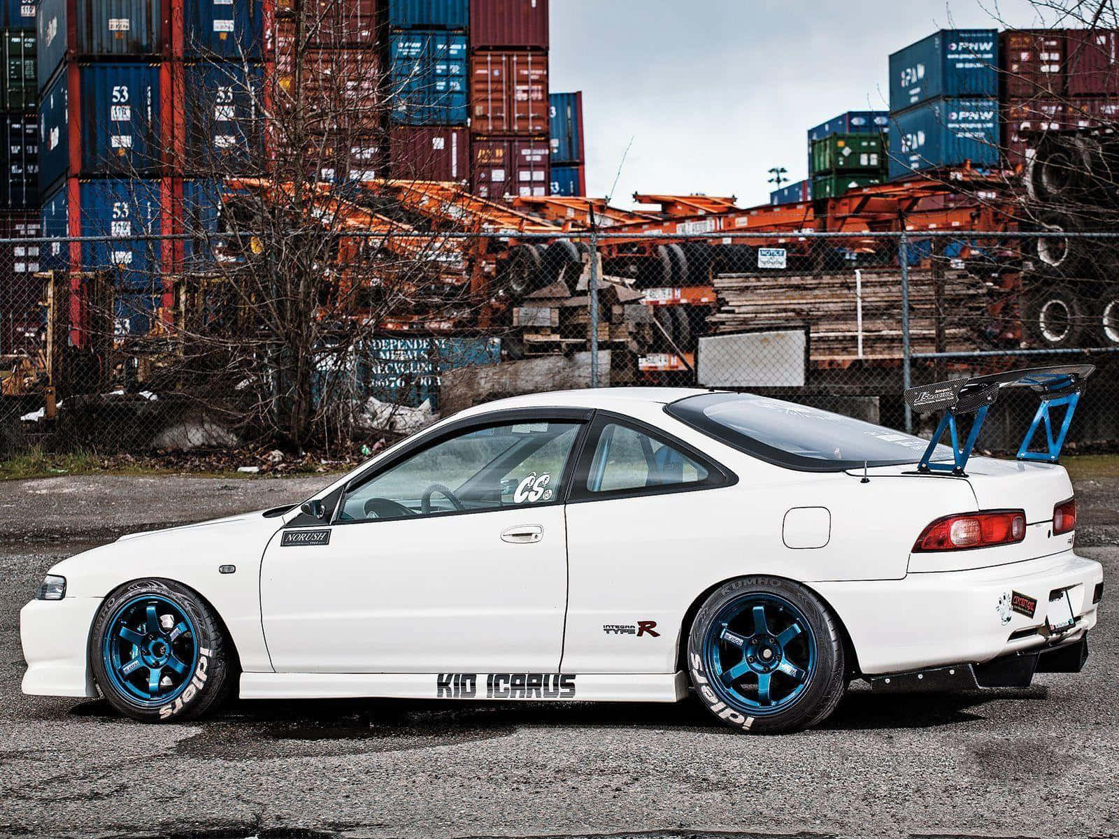 Acura Integra 桌布 壁紙 — 一輛白色改裝跑車配藍色輪圈，停泊在工業區堆疊的貨運集裝箱前。