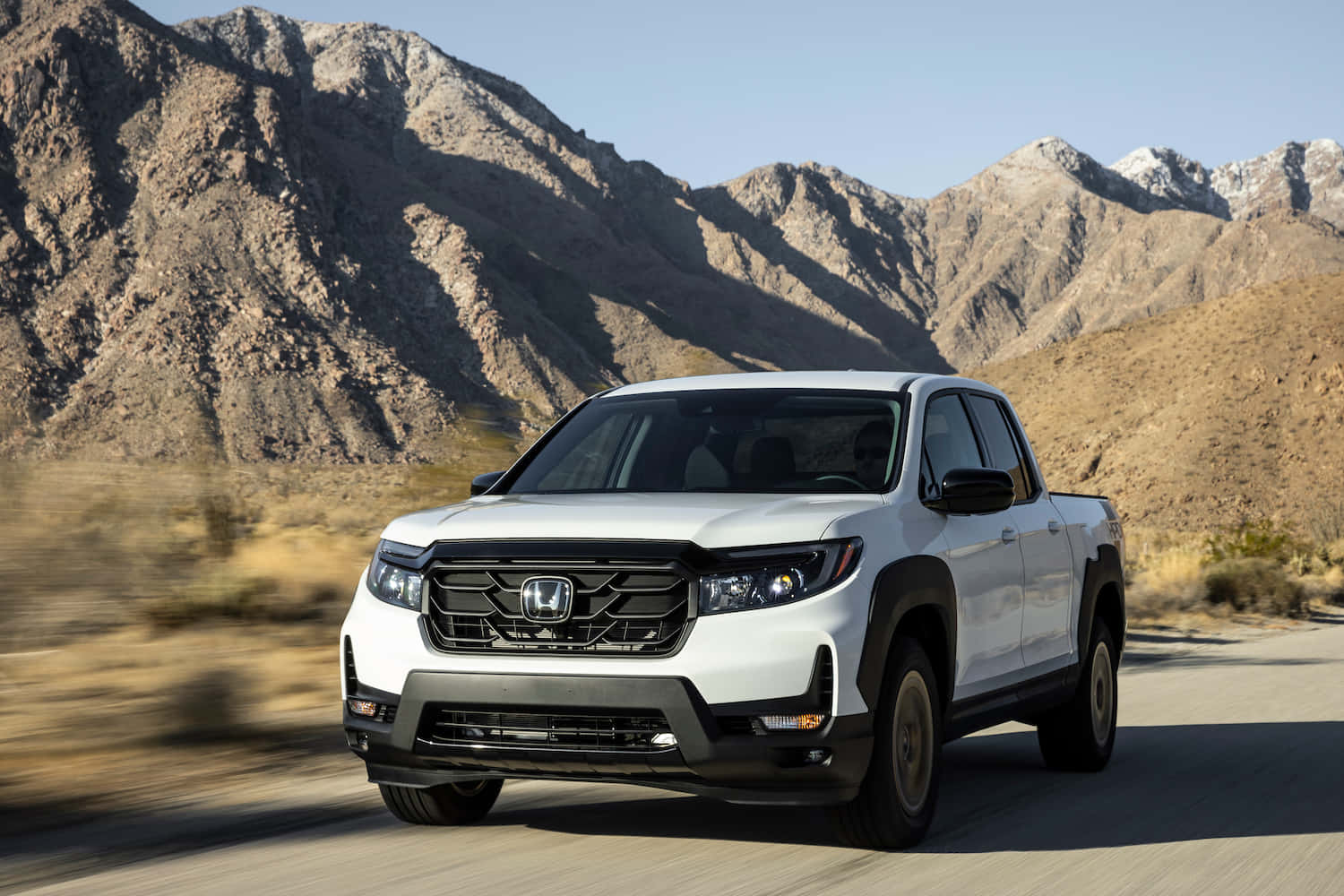 Honda Ridgeline 桌布 壁紙 — 一輛白色本田Ridgeline皮卡車在晴朗藍天下，沿著沙漠公路疾馳，背景是崎嶇的山脈。