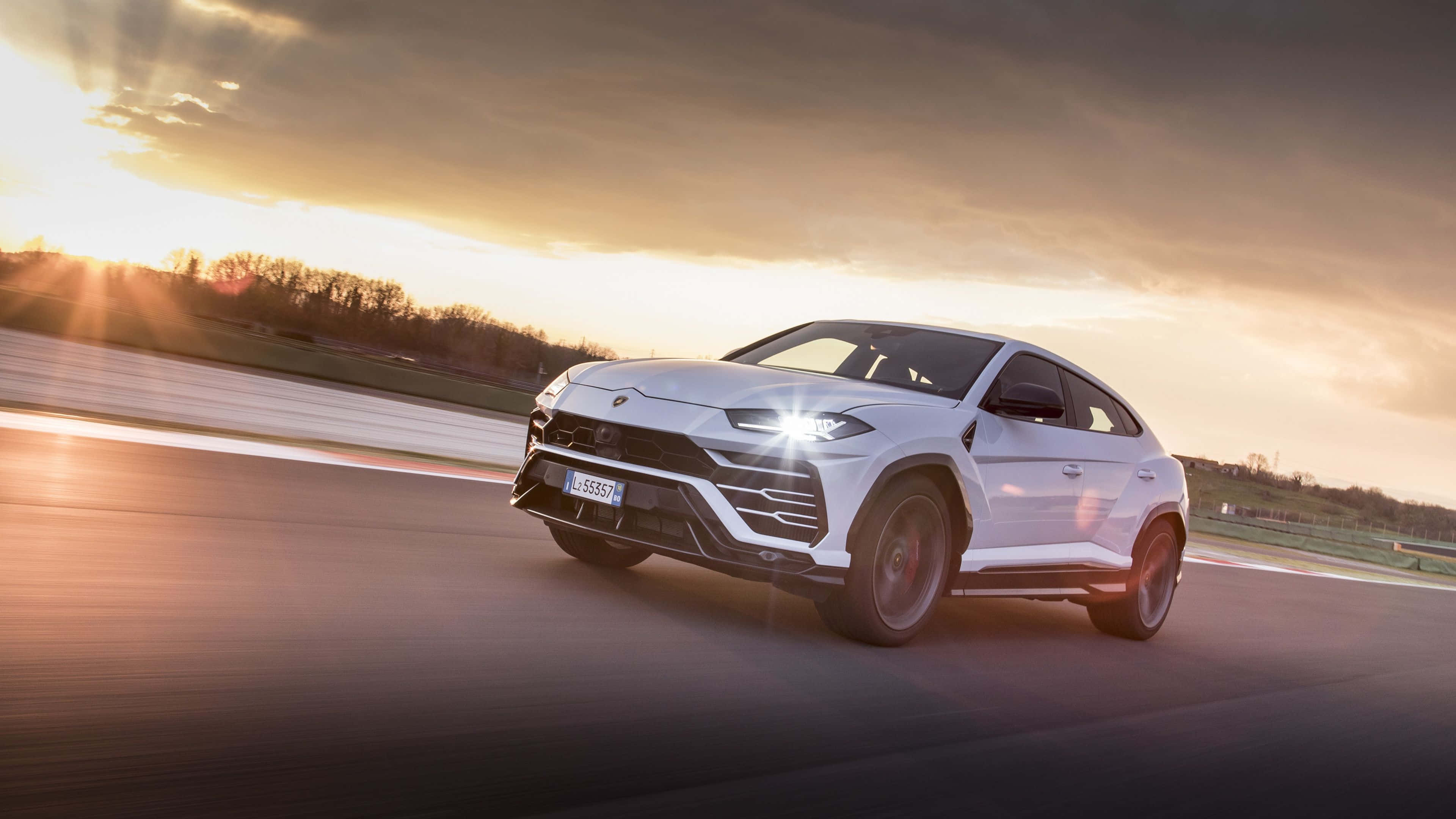 Lamborghini Urus 桌布 壁紙 — 一輛白色林寶堅尼 Urus 在日落時分沿著賽道疾馳，帶有戲劇性的鏡頭光暈和動態模糊效果。