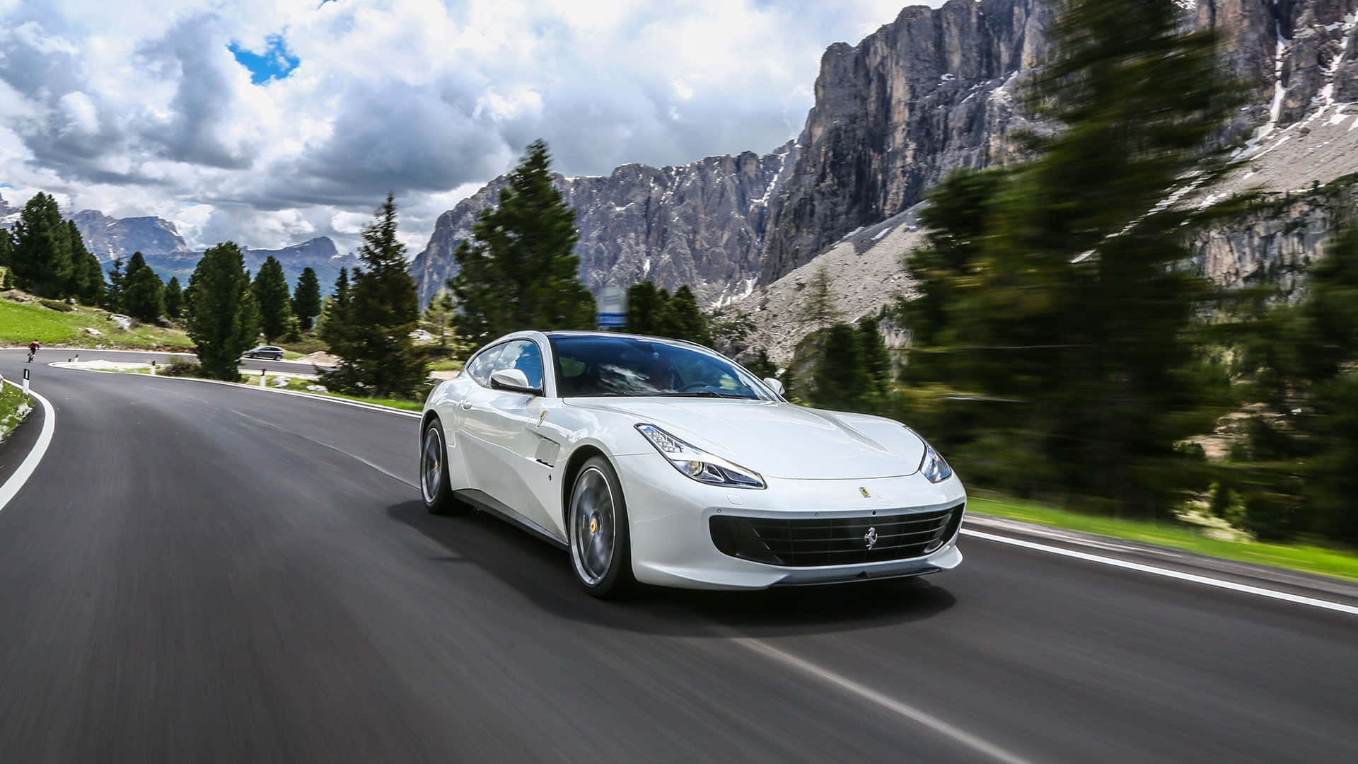 Ferrari Gtc4lusso 桌布 壁紙 — 一輛白色法拉利在蜿蜒的山路上快速行駛，兩旁是茂密的綠樹和崎嶇的山峰，天空多雲。