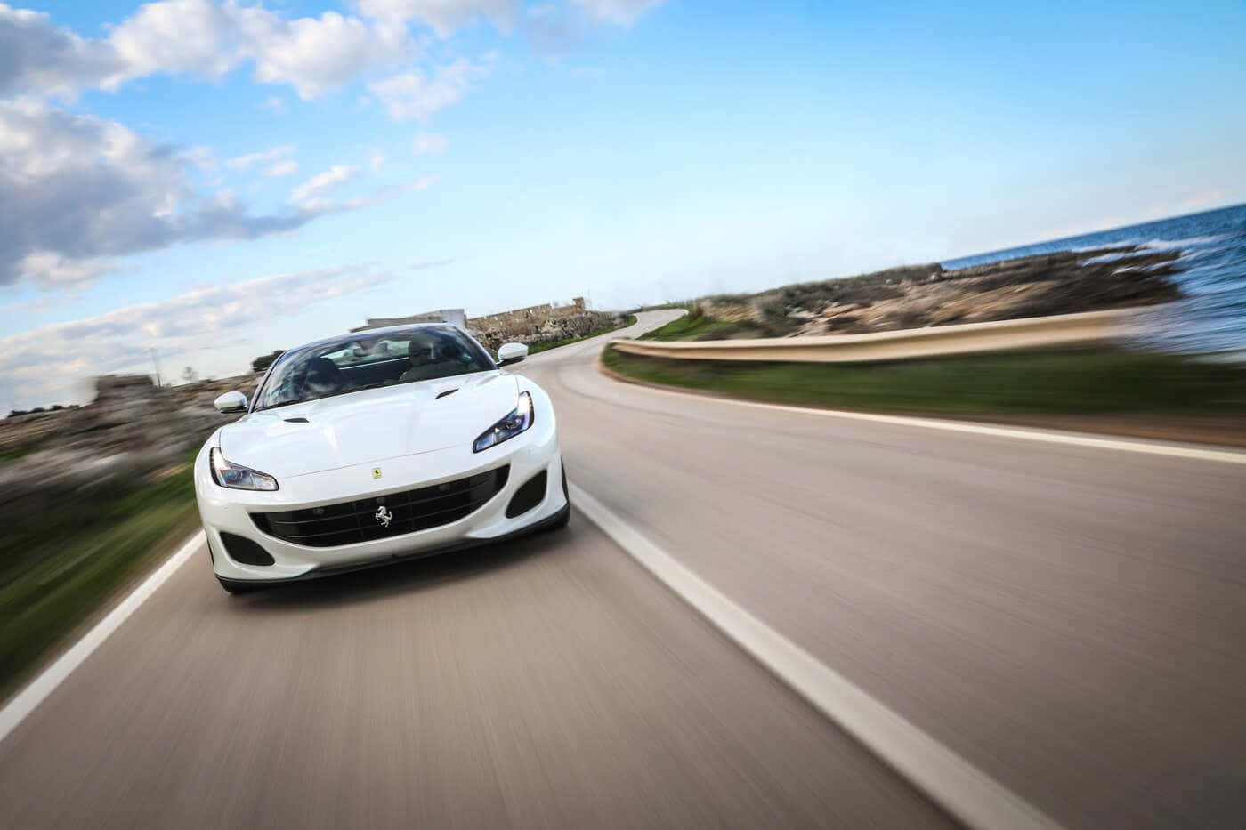 Ferrari Portofino 桌布 壁紙 — 一輛白色法拉利跑車沿著海岸公路疾馳，在藍天和海洋的背景下呈現模糊的動態效果。
