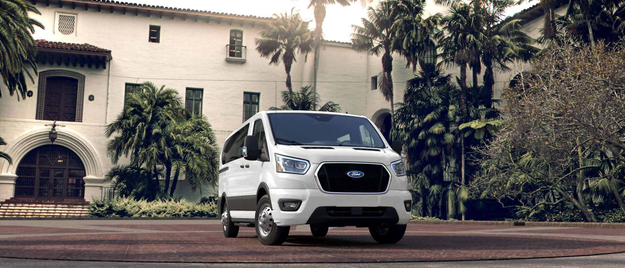 Ford Transit 桌布 壁紙 — 一輛白色福特 Transit 客貨車停泊在磚砌車道上，前方是西班牙風格建築，周圍環繞著棕櫚樹。