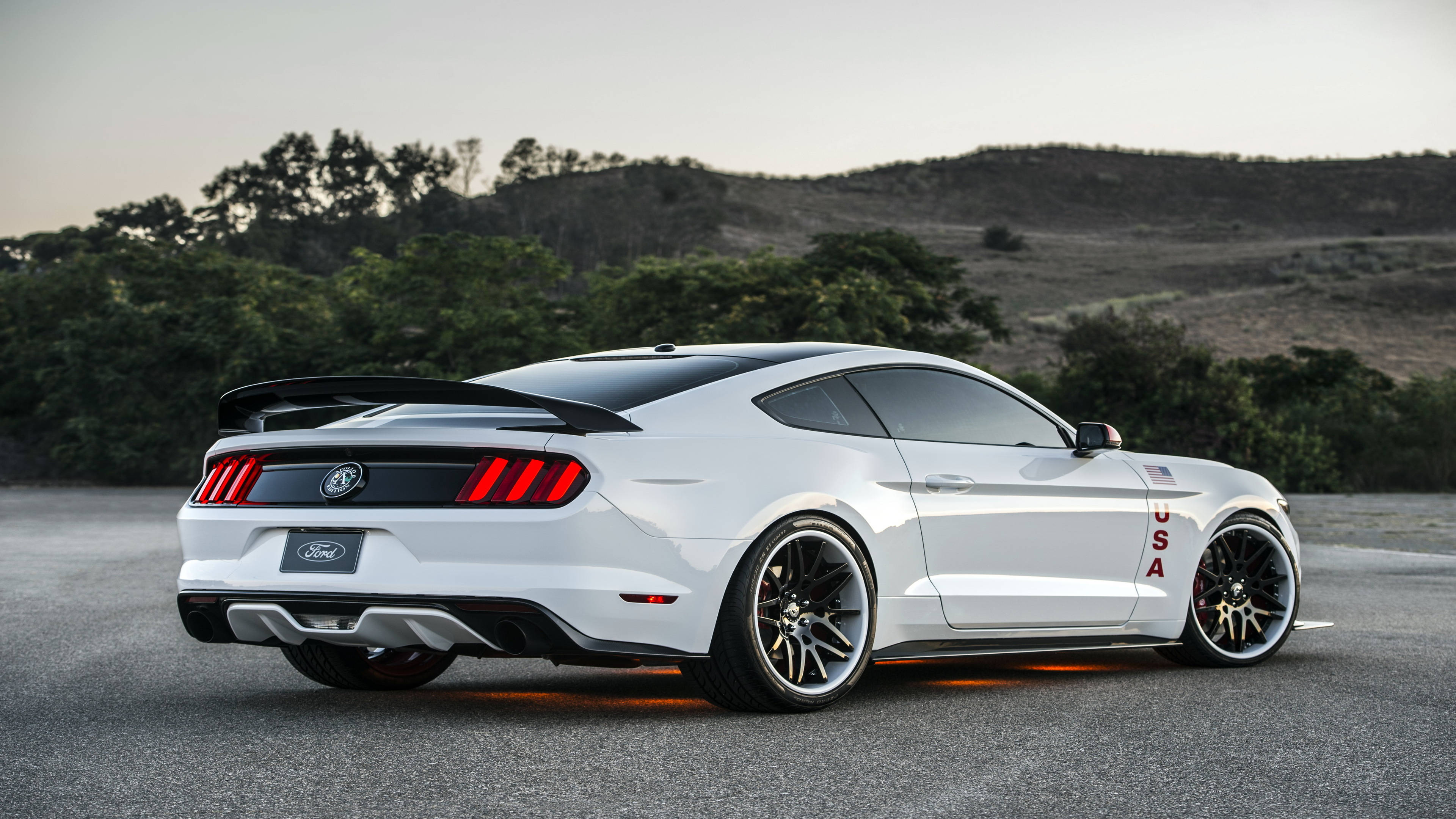 mustang HD 桌布 壁紙 — 一輛白色福特野馬，帶有紅色尾燈和美國貼紙，停泊在瀝青路上，背景是黃昏時分的丘陵景觀。