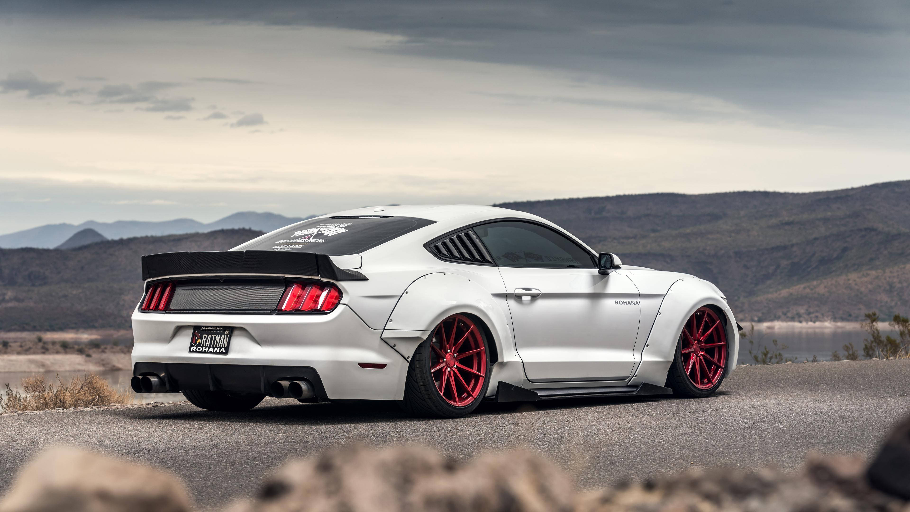 mustang HD 桌布 壁紙 — 一輛白色福特野馬配紅色輪胎，停泊在沙漠公路上，背景有山脈和湖泊，天空多雲。