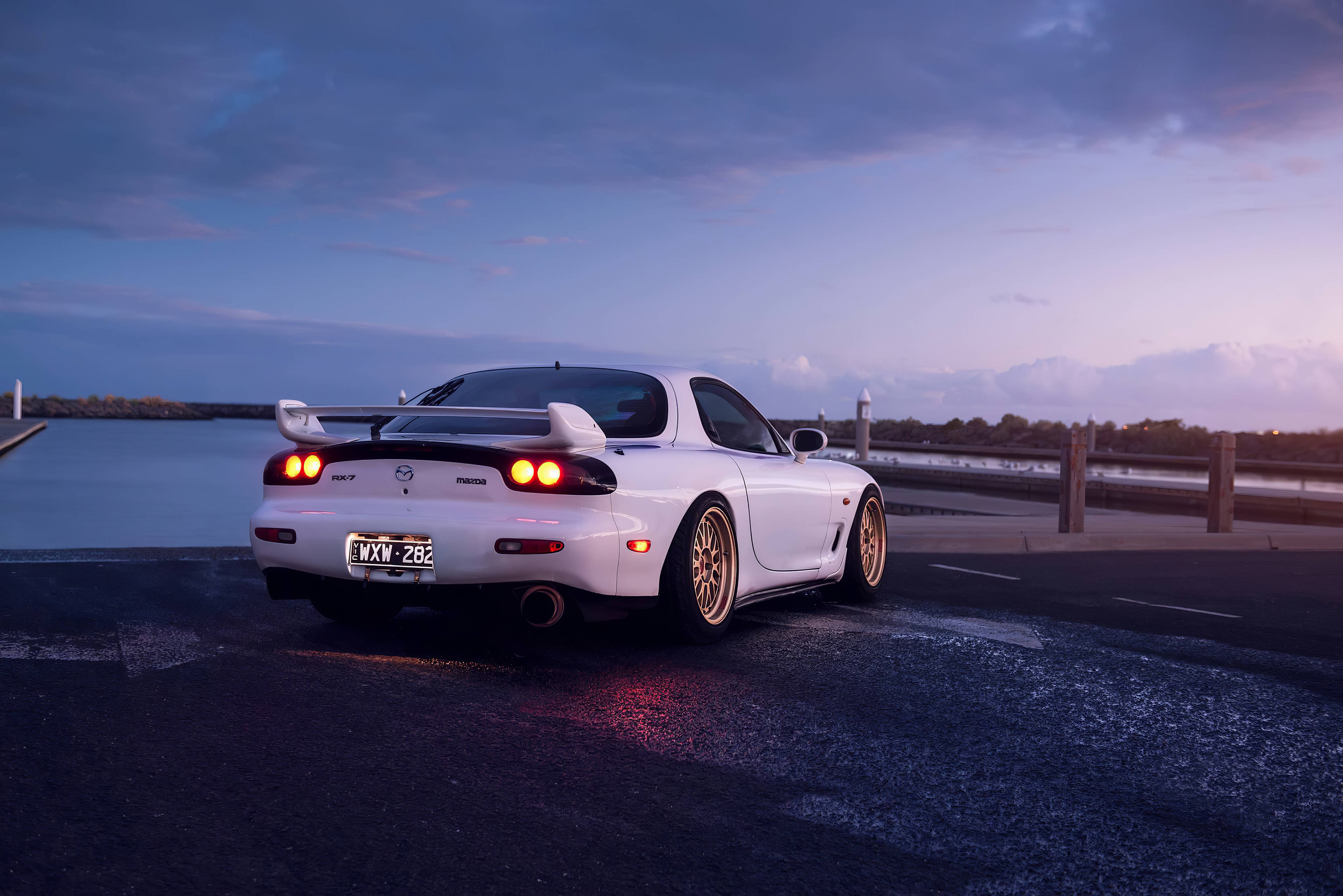 Mazda 桌布 壁紙 — 一輛白色萬事得RX-7跑車配金色輪圈，在紫色天空下，於平靜水邊的濕滑道路上，在黃昏中閃耀。