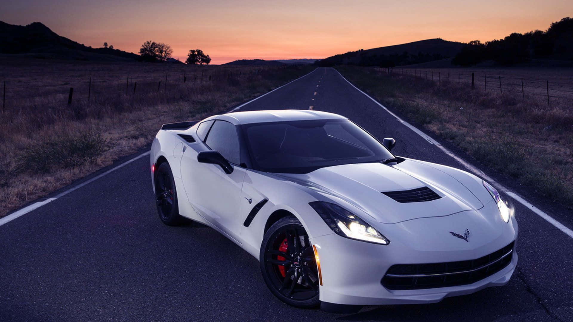 Chevrolet Corvette Z06 桌布 壁紙 — 一輛白色跑車亮著車頭燈，在日落時分行駛在一條漫長的鄉村道路上，背景是山丘。