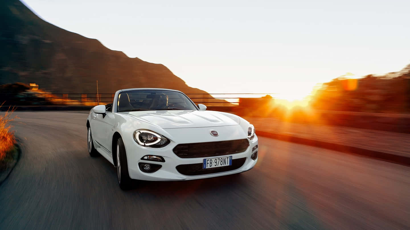 Fiat 124 Spider 桌布 壁紙 — 一輛白色開篷跑車在日落時分沿著蜿蜒的山路飛馳，身後閃耀著金色的光芒。