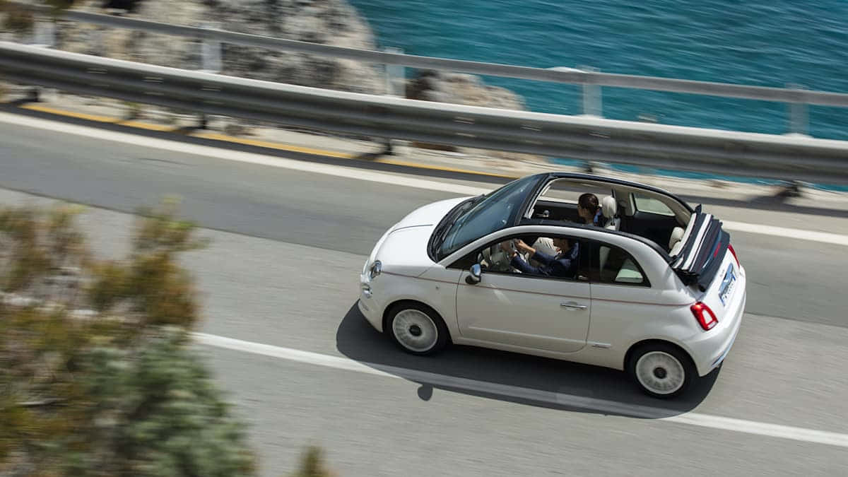 Fiat 500 桌布 壁紙 — 一輛白色開篷車沿著海岸公路行駛，背景是碧綠色的海洋和岩石峭壁。