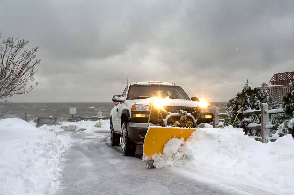 Snowplow 桌布 壁紙 — 一輛白色除雪車在灰色冬日天空下清理一條被雪覆蓋的狹窄沿海道路。