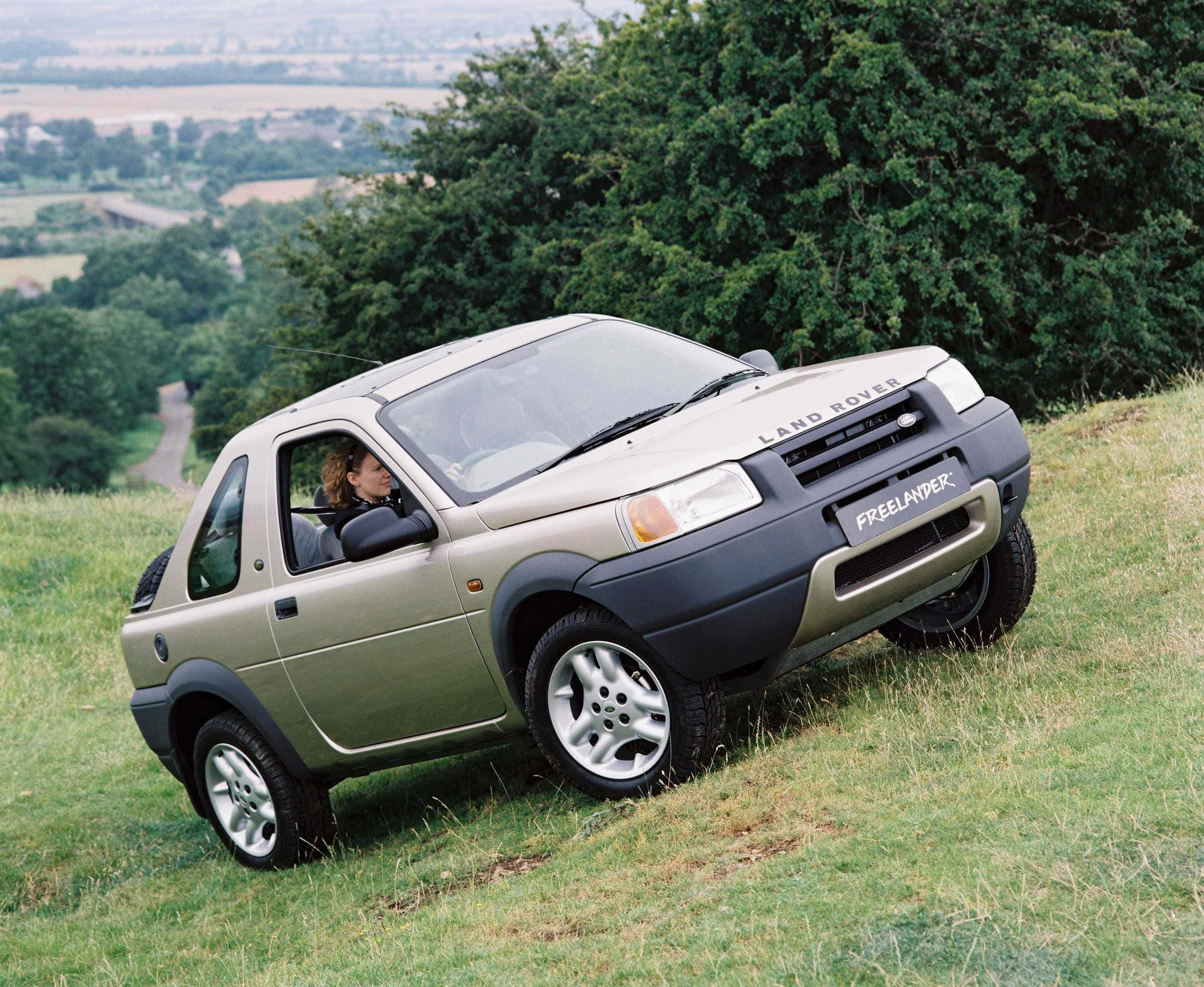 Land Rover Freelander 桌布 壁紙 — 一輛米色Land Rover Freelander停泊在長滿草的山坡上，車內有一位女士，俯瞰著鬱鬱蔥蔥的綠色山谷和遠處的樹木。