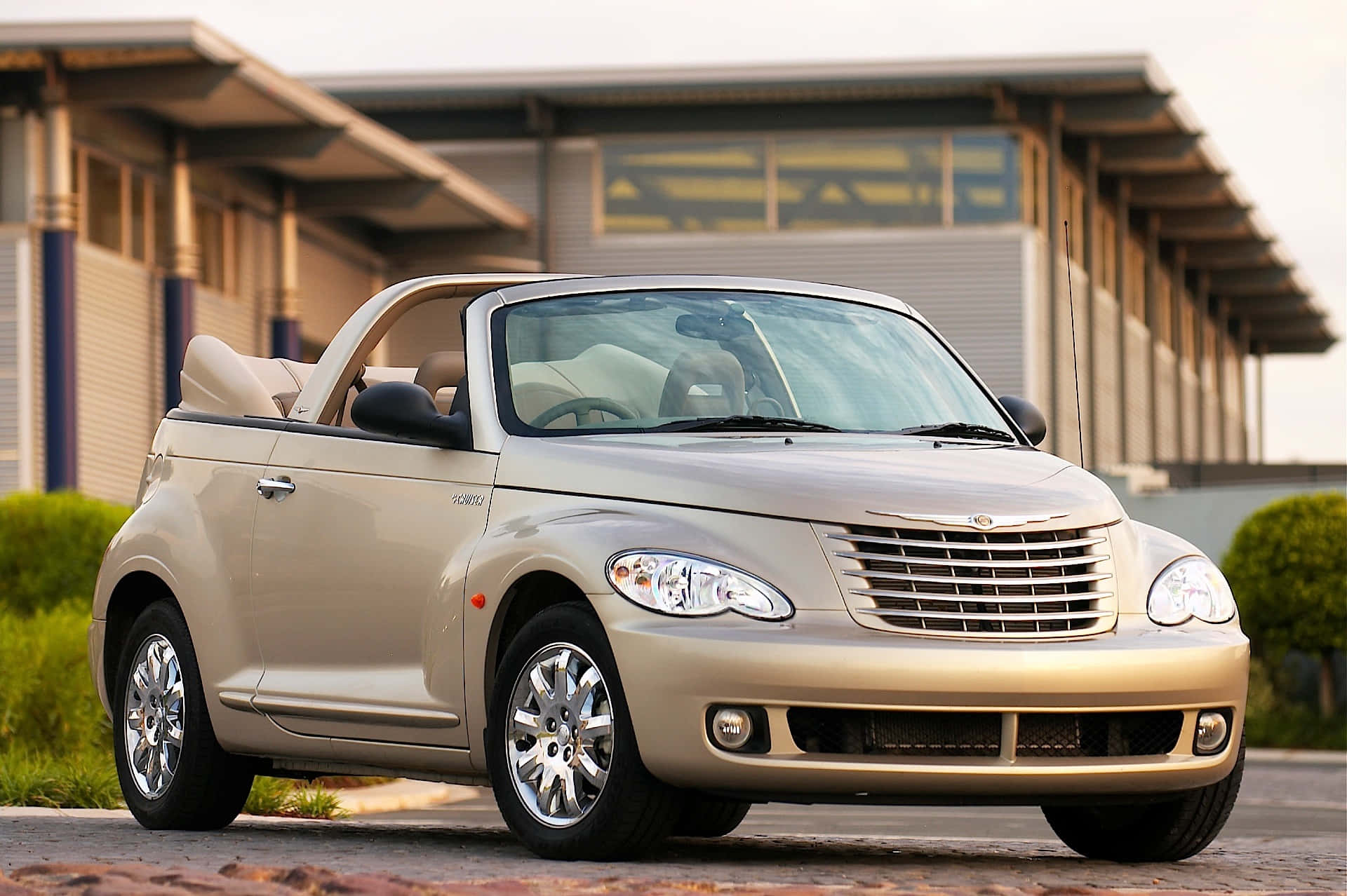 Chrysler Pt Cruiser 桌布 壁紙 — 一輛米色克萊斯勒 PT Cruiser 敞篷車停泊在磚砌車道上，背景是現代建築和綠色灌木。