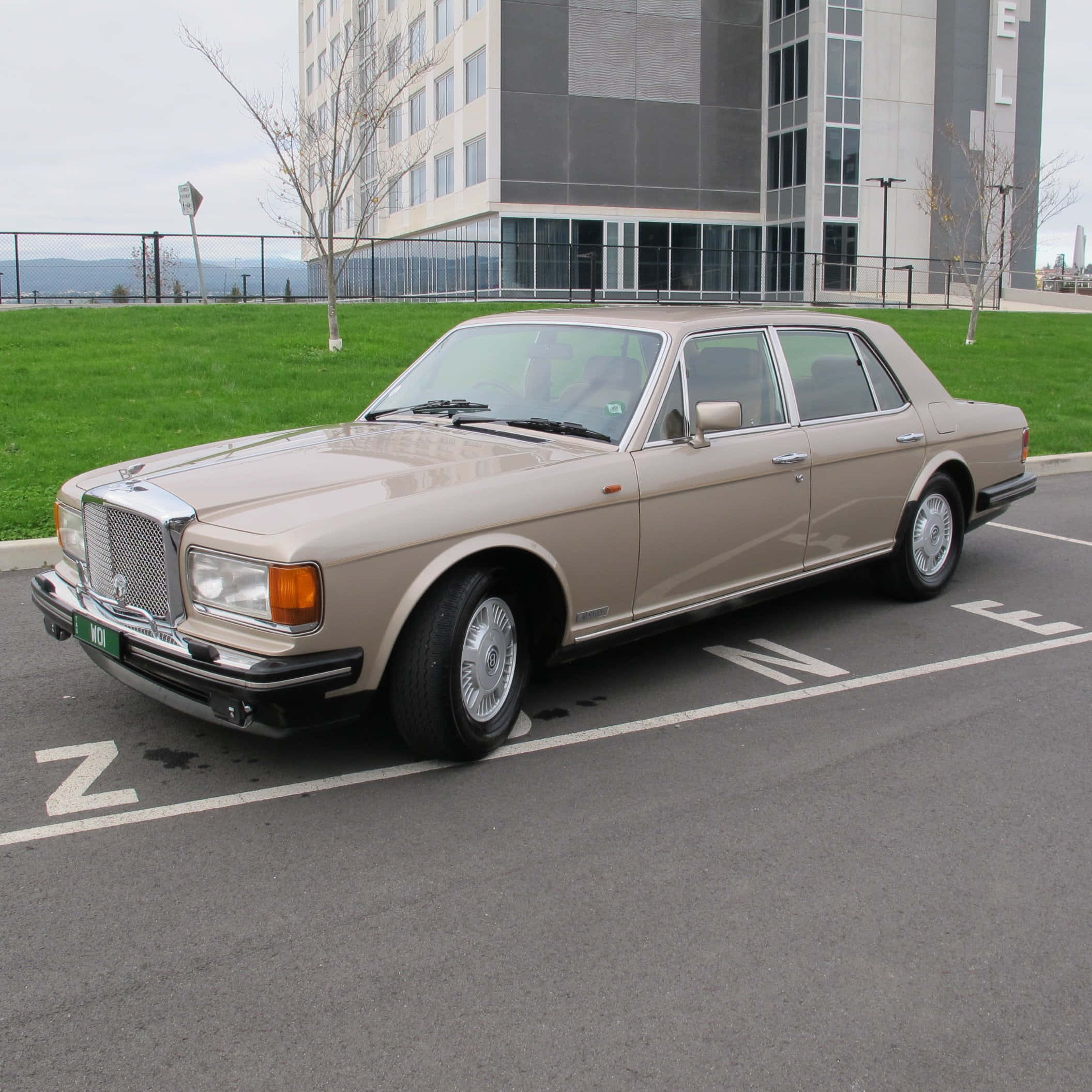 Bentley Eight 桌布 壁紙 — 一輛米色勞斯萊斯銀靈停泊在現代建築旁邊的標記車位，建築物周圍有綠色草地和光禿禿的樹木，天空多雲。