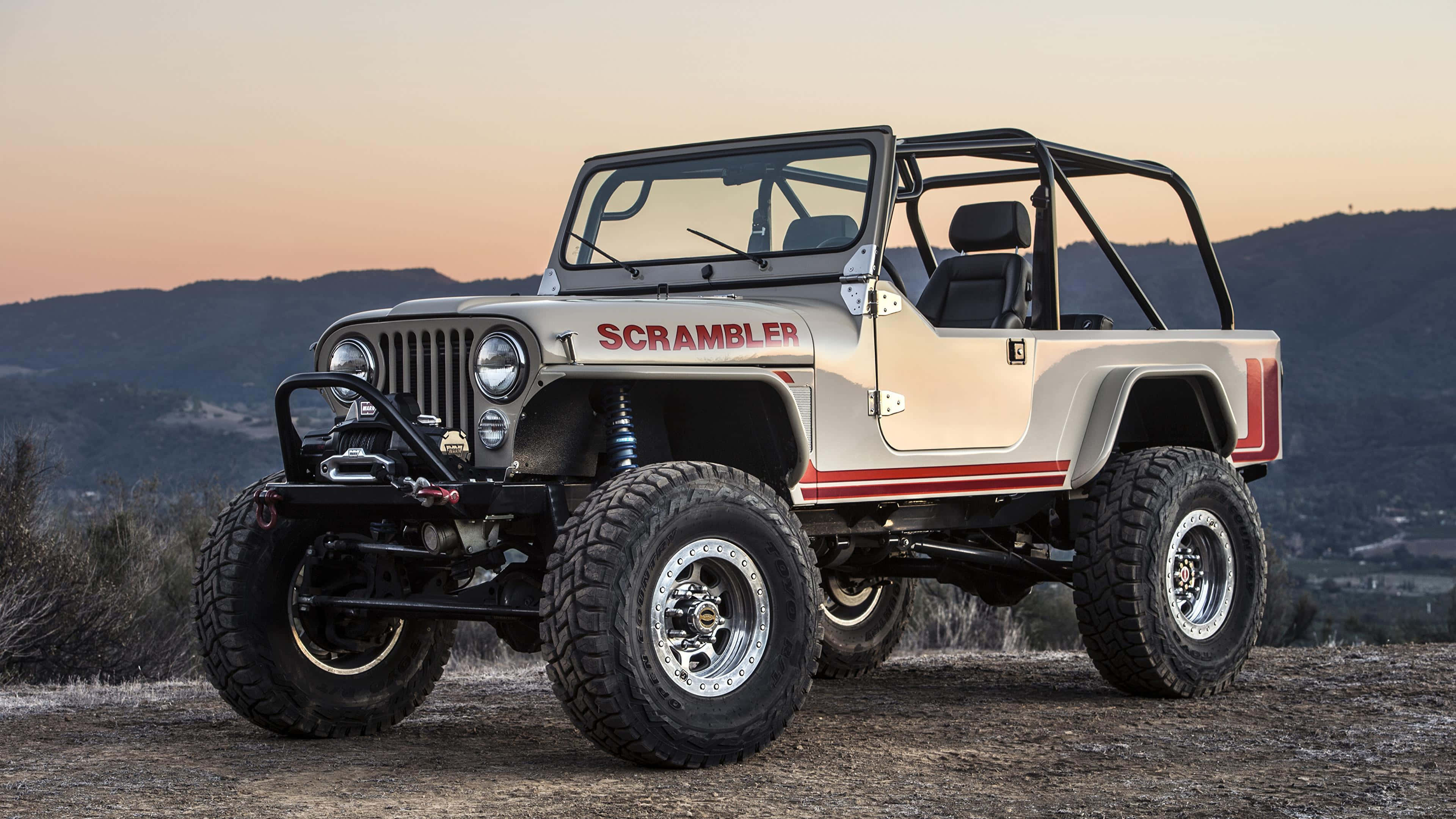 Jeep Cj 桌布 壁紙 — 一輛米色吉普Scrambler帶有紅色條紋和超大輪胎，在日落時分站在土丘上，背景是山脈。